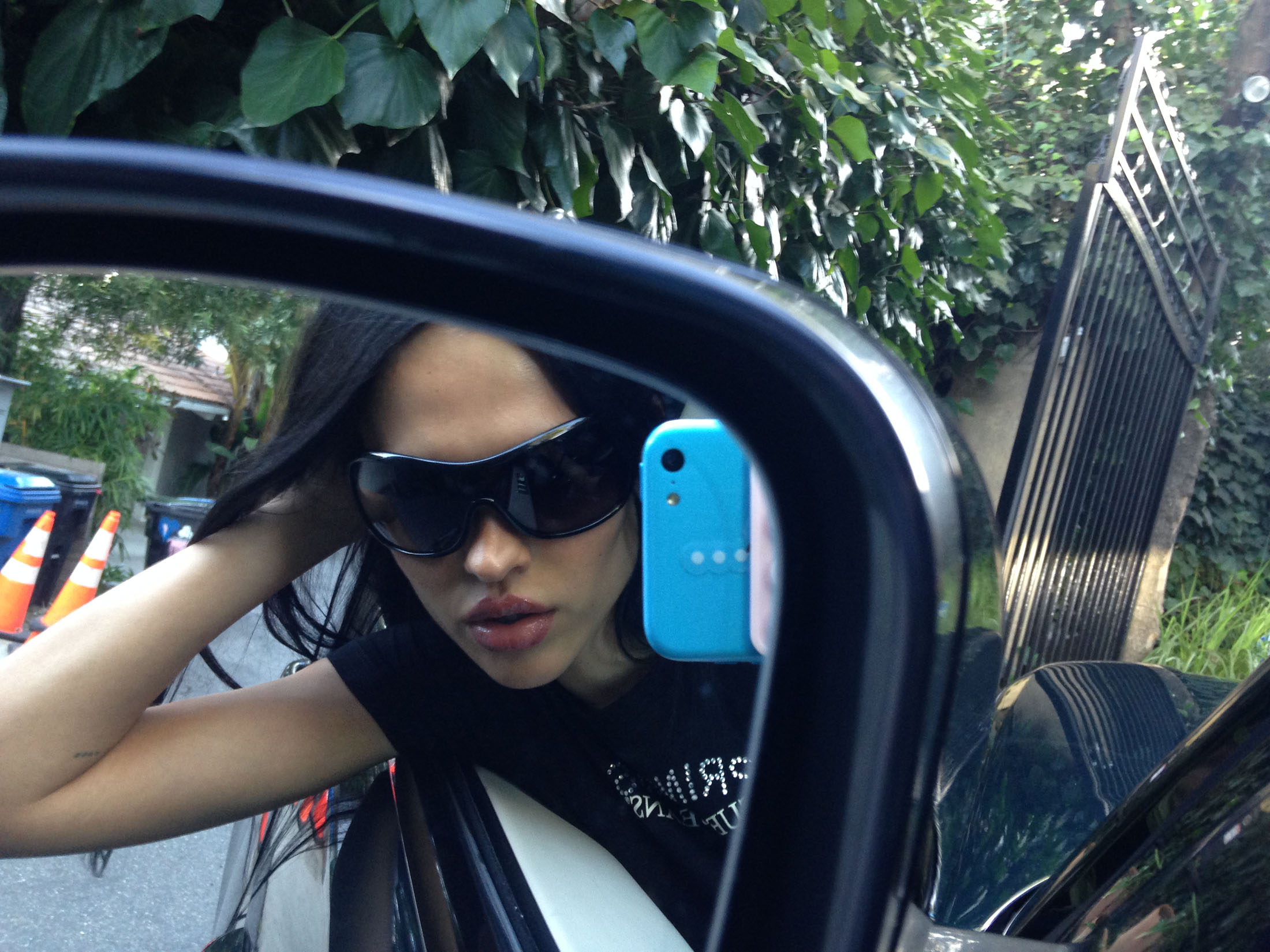 Brunette woman takes a selfie in the side mirror of a car, showcasing sunglasses and a casual outfit, with leafy outdoor background.