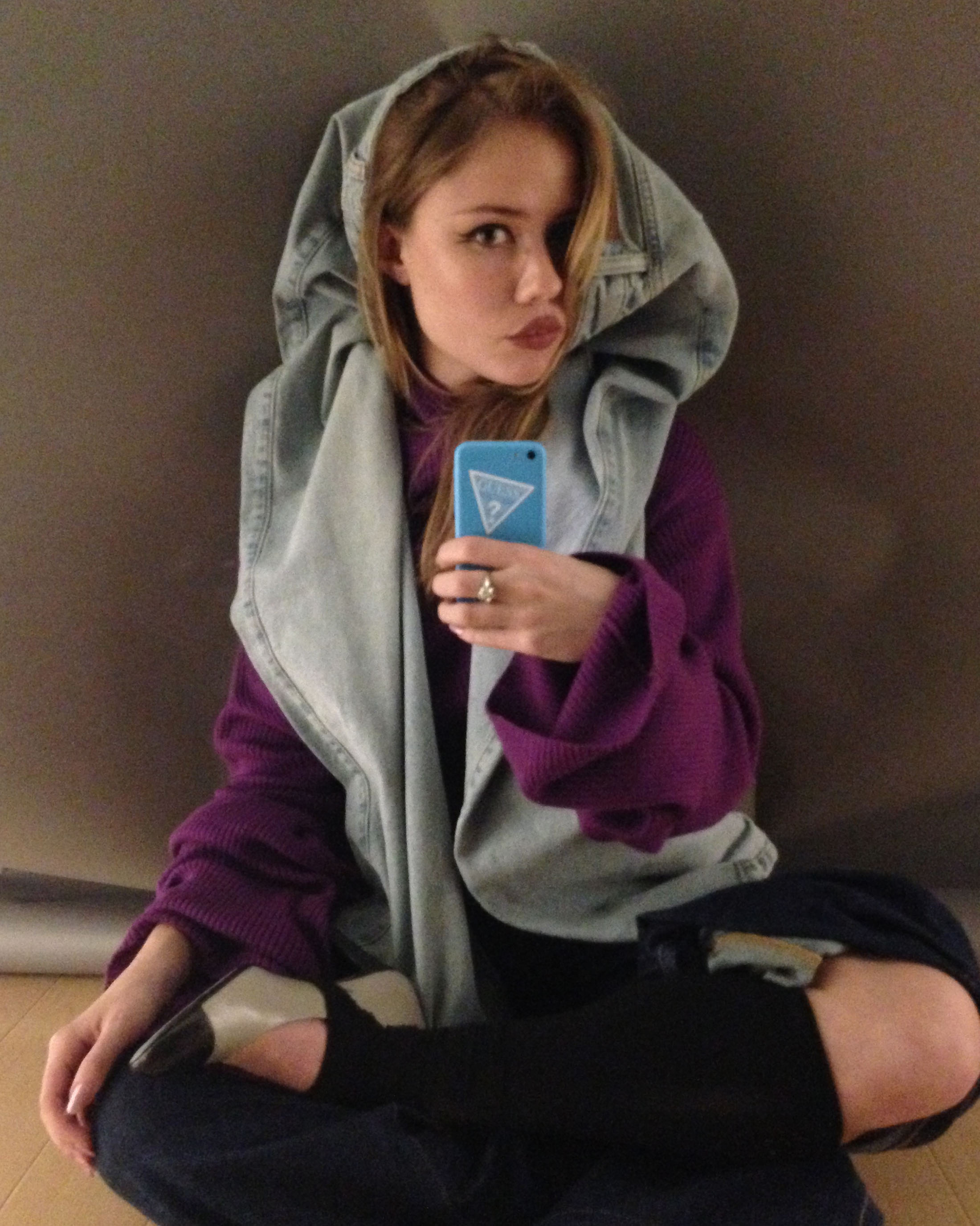 Young adult woman in a purple sweater and jeans poses for a selfie indoors with a blue phone, wearing a denim hooded wrap.