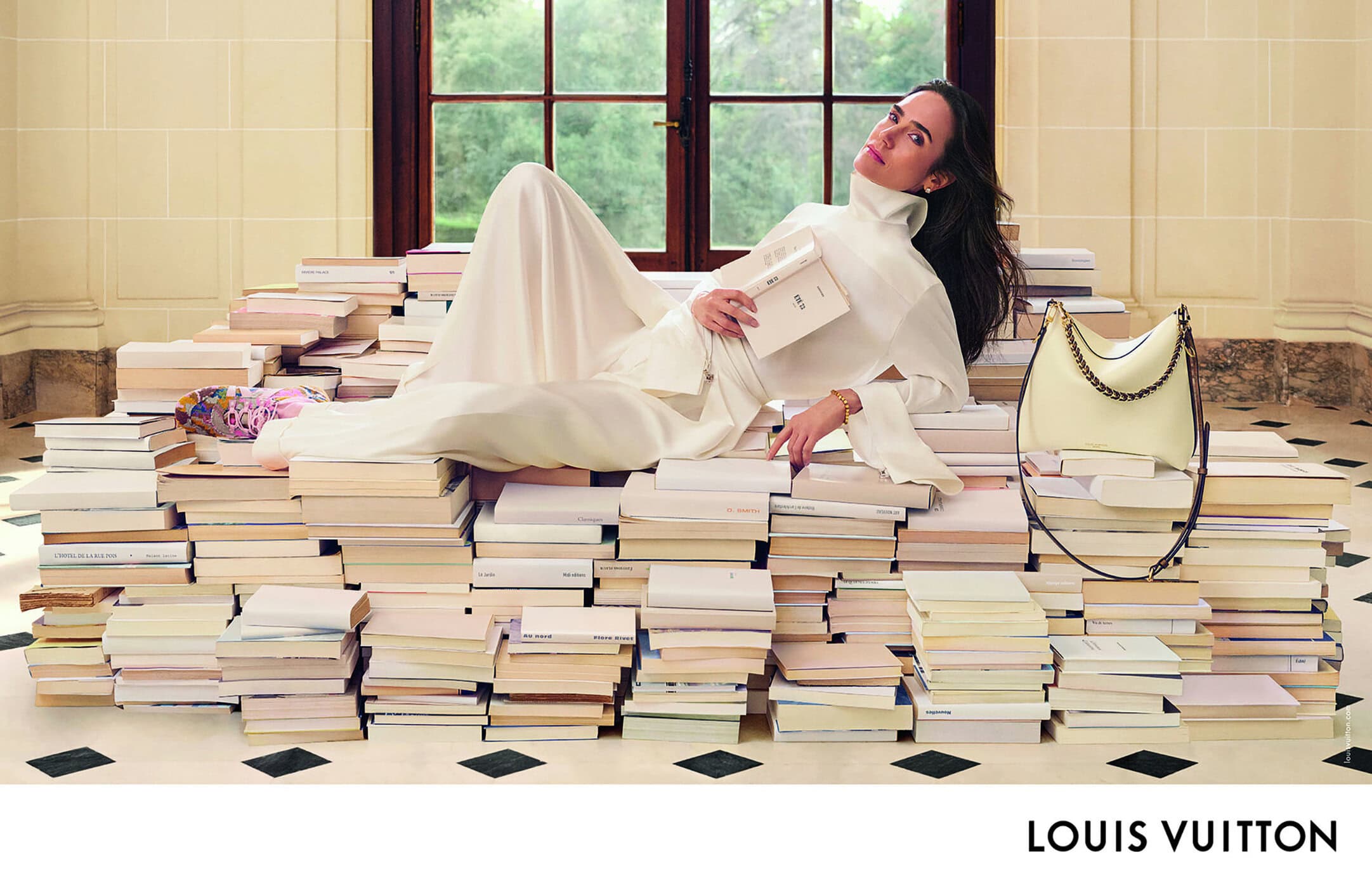 Jennifer Connelly reclines on a book stack in a cream dress, holding a book, with a cream Louis Vuitton bag in an indoor setting.