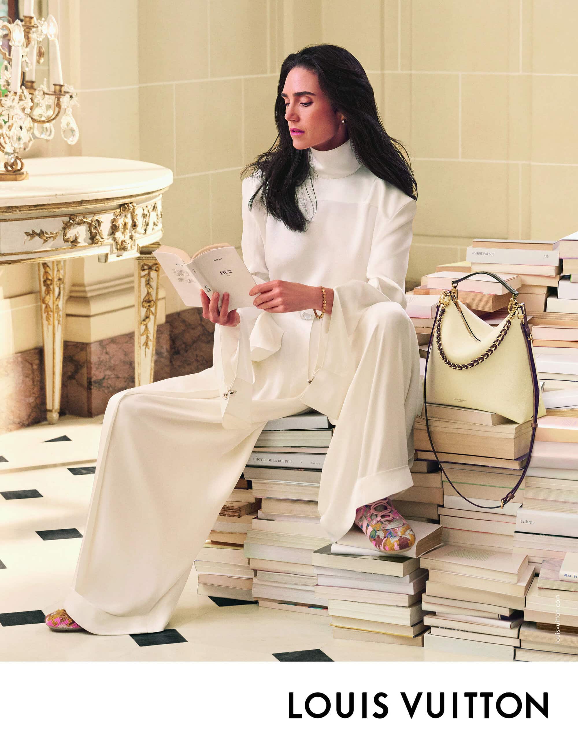 Fashion shot of a woman in a white outfit seated on books and reading in an indoor environment next to a side table and handbag.