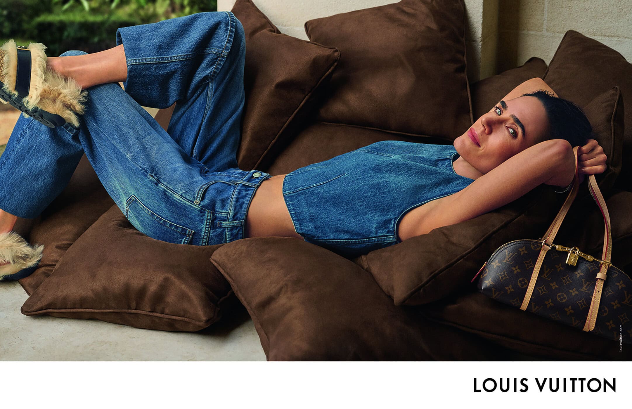 Woman lying on brown pillows in a blue denim outfit with a Louis Vuitton handbag, soft lighting, and a relaxed pose.