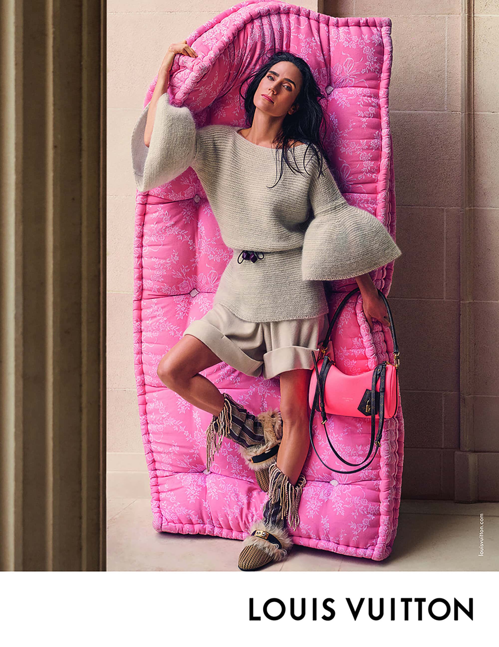 Jennifer Connelly poses with a pink tufted chaise lounge and pink purse in a fashion editorial shot in a neutral indoor setting.