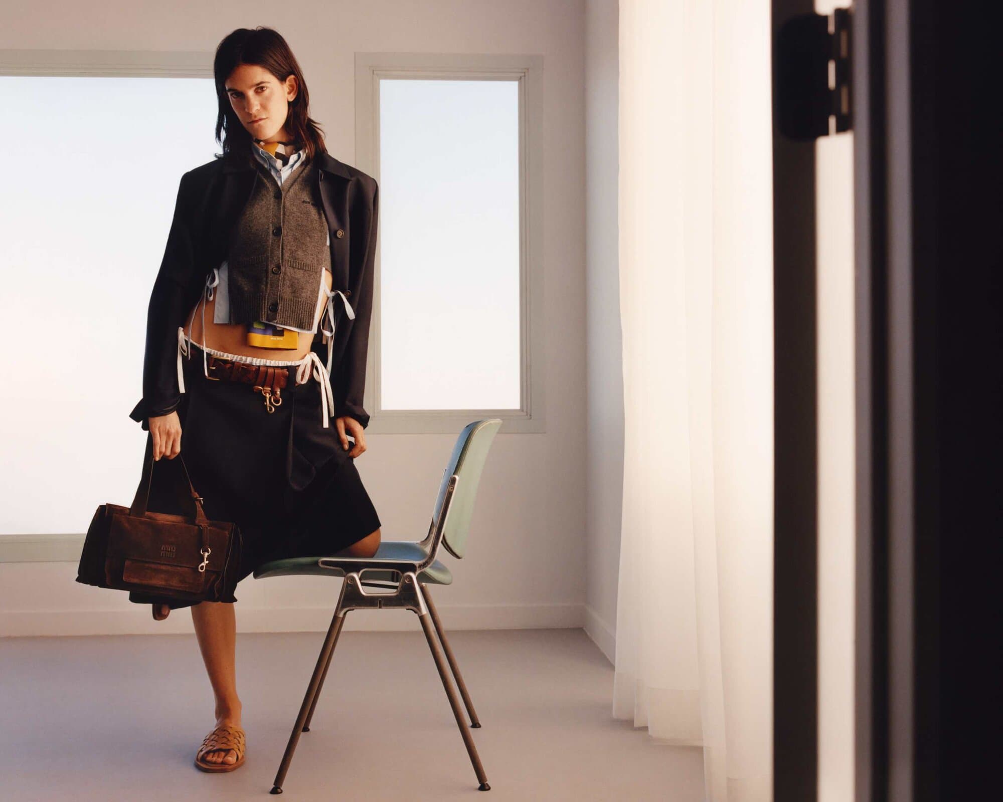 A woman in a black skirt and jacket, tan sandals, and carrying a brown leather handbag is sitting on a metal frame chair indoors.
