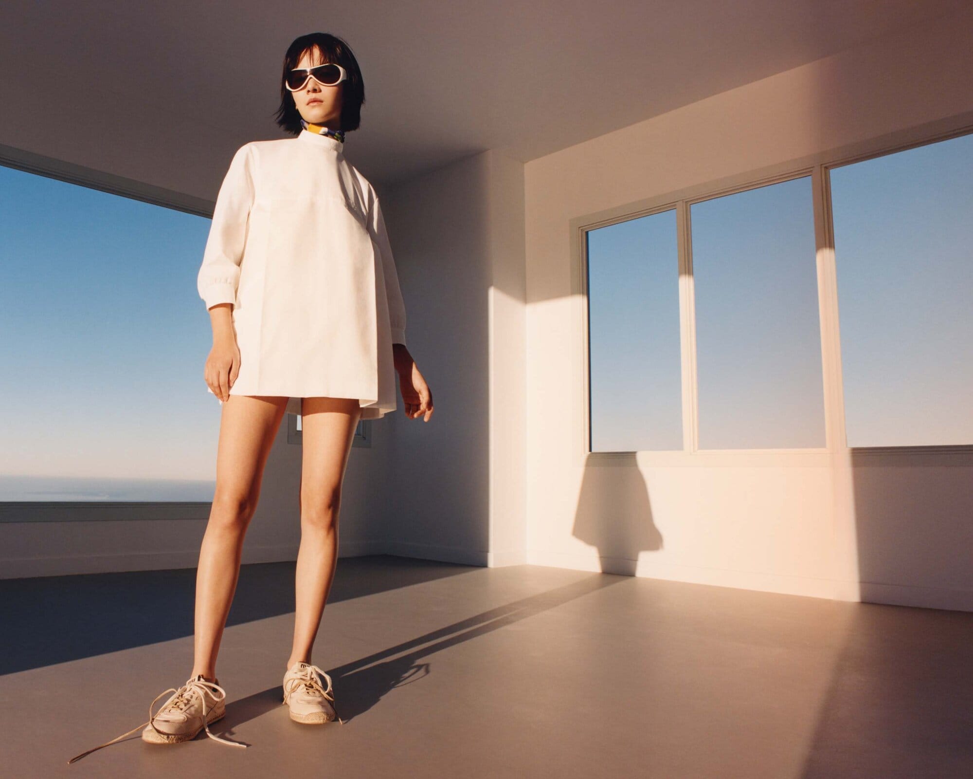 A slender woman poses in a minimalist white dress, white sneakers and sunglasses, standing in an empty white room with windows.