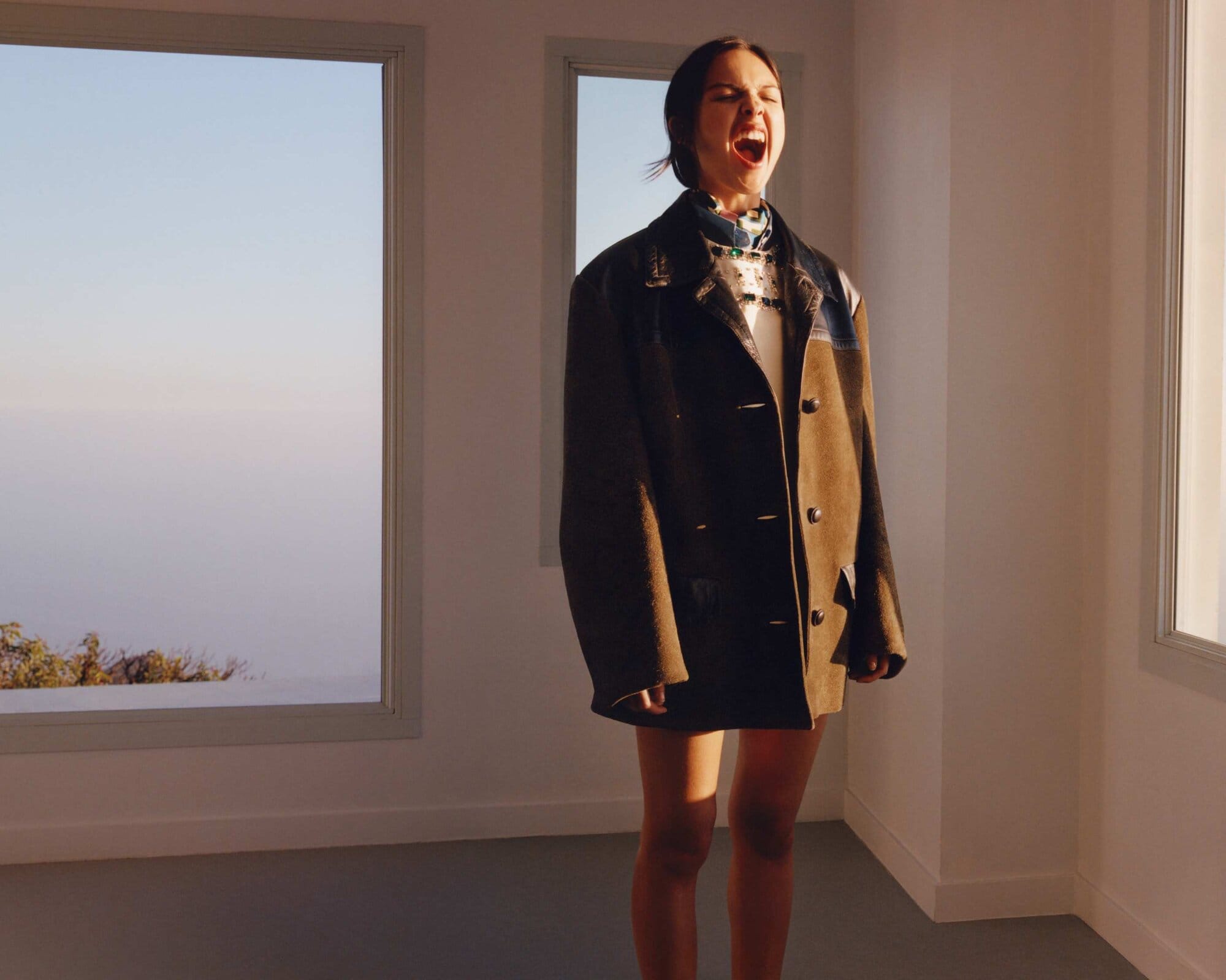 Young woman in an oversized brown coat stands in a brightly lit room with large windows framing a blue sky and scenic outdoor vista.