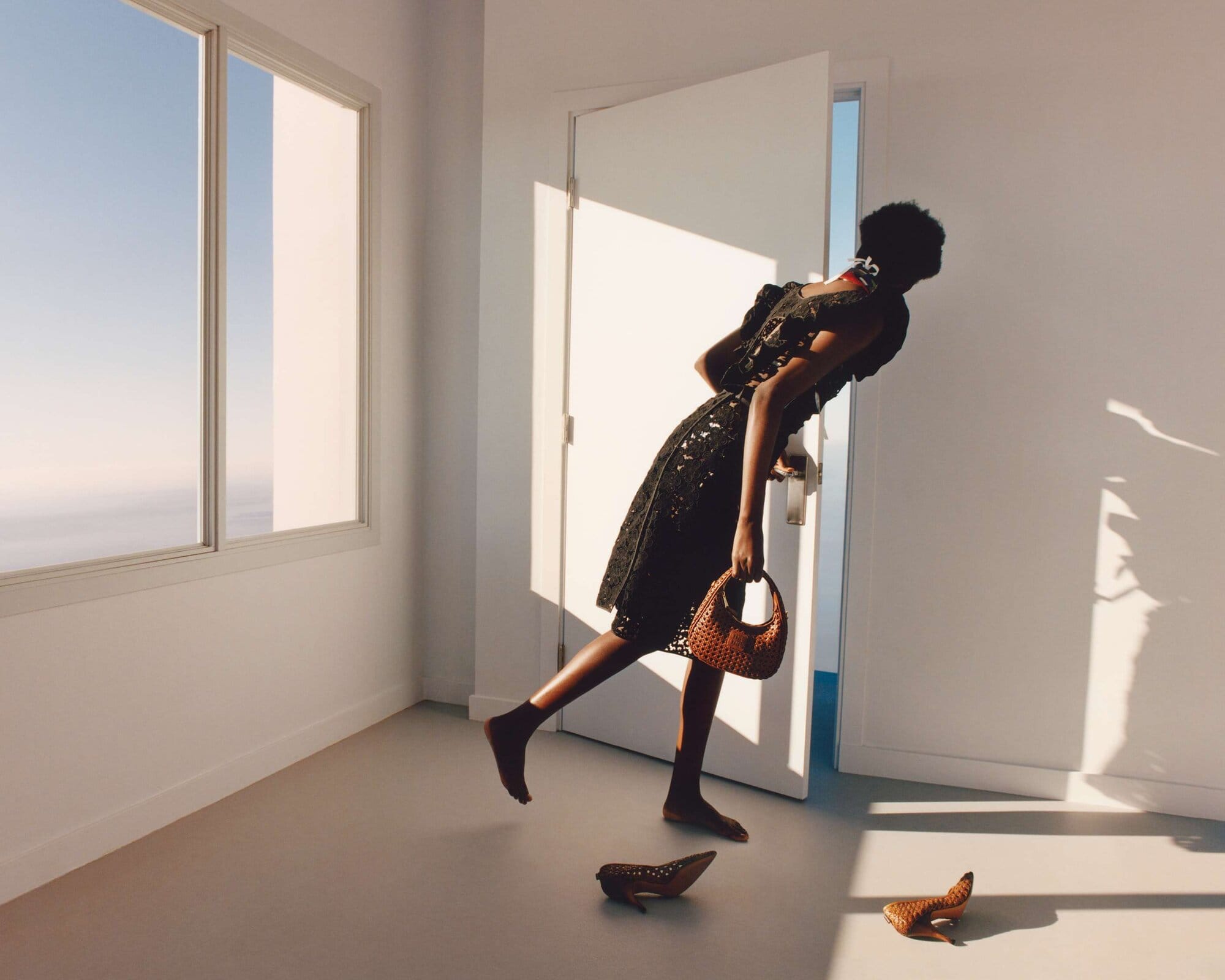 A woman in a black dress enters a bright white room, carrying a brown woven purse. Shoes are on the floor near an open door and window.