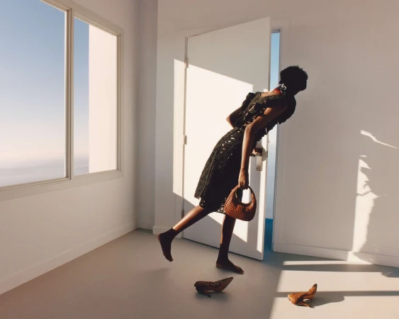 A woman in a black dress enters a bright white room, carrying a brown woven purse. Shoes are on the floor near an open door and window.