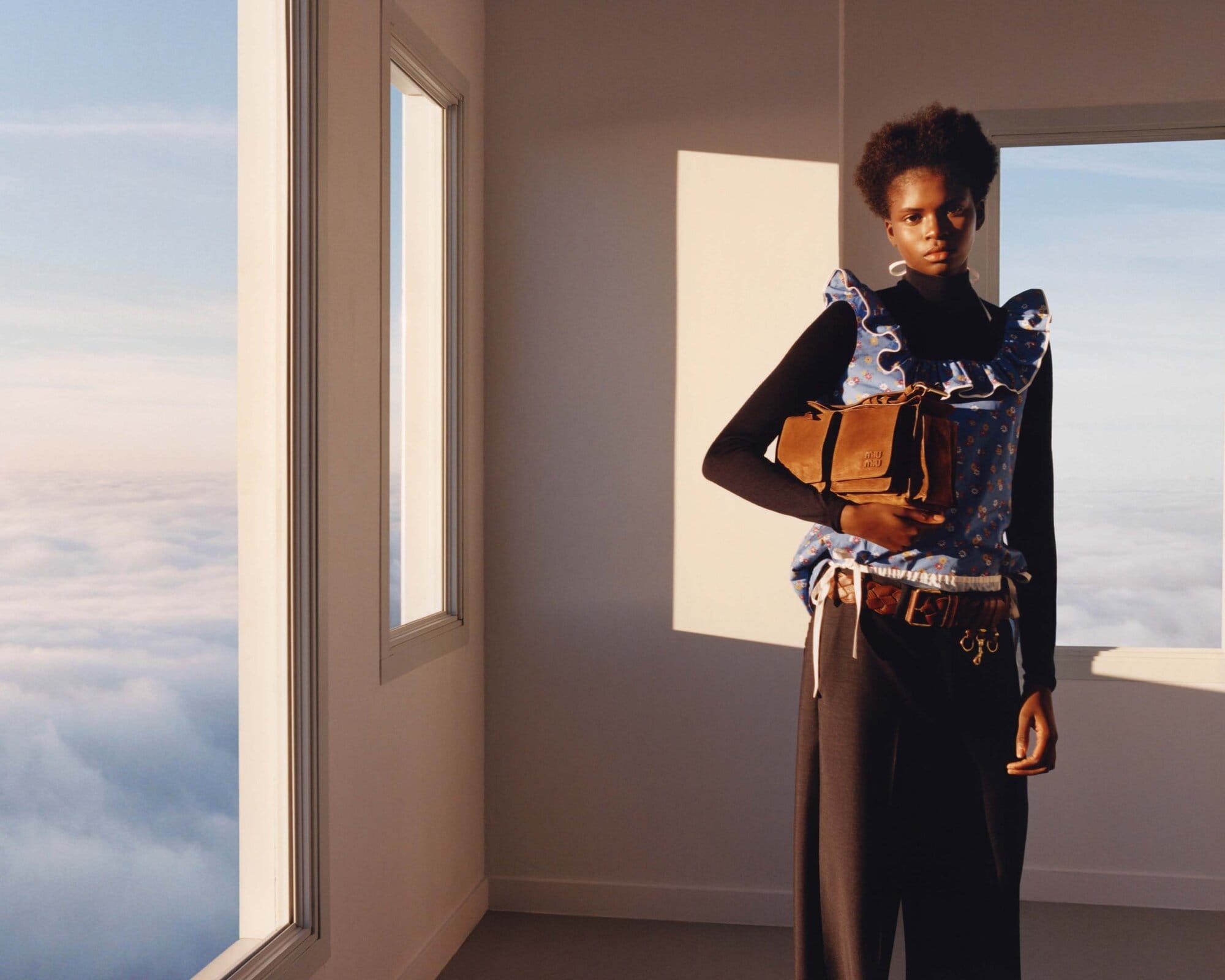 An African-American woman in a blue floral top holds a brown handbag, posing in an interior with large windows and a cloudscape view.