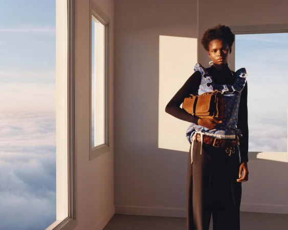 An African-American woman in a blue floral top holds a brown handbag, posing in an interior with large windows and a cloudscape view.