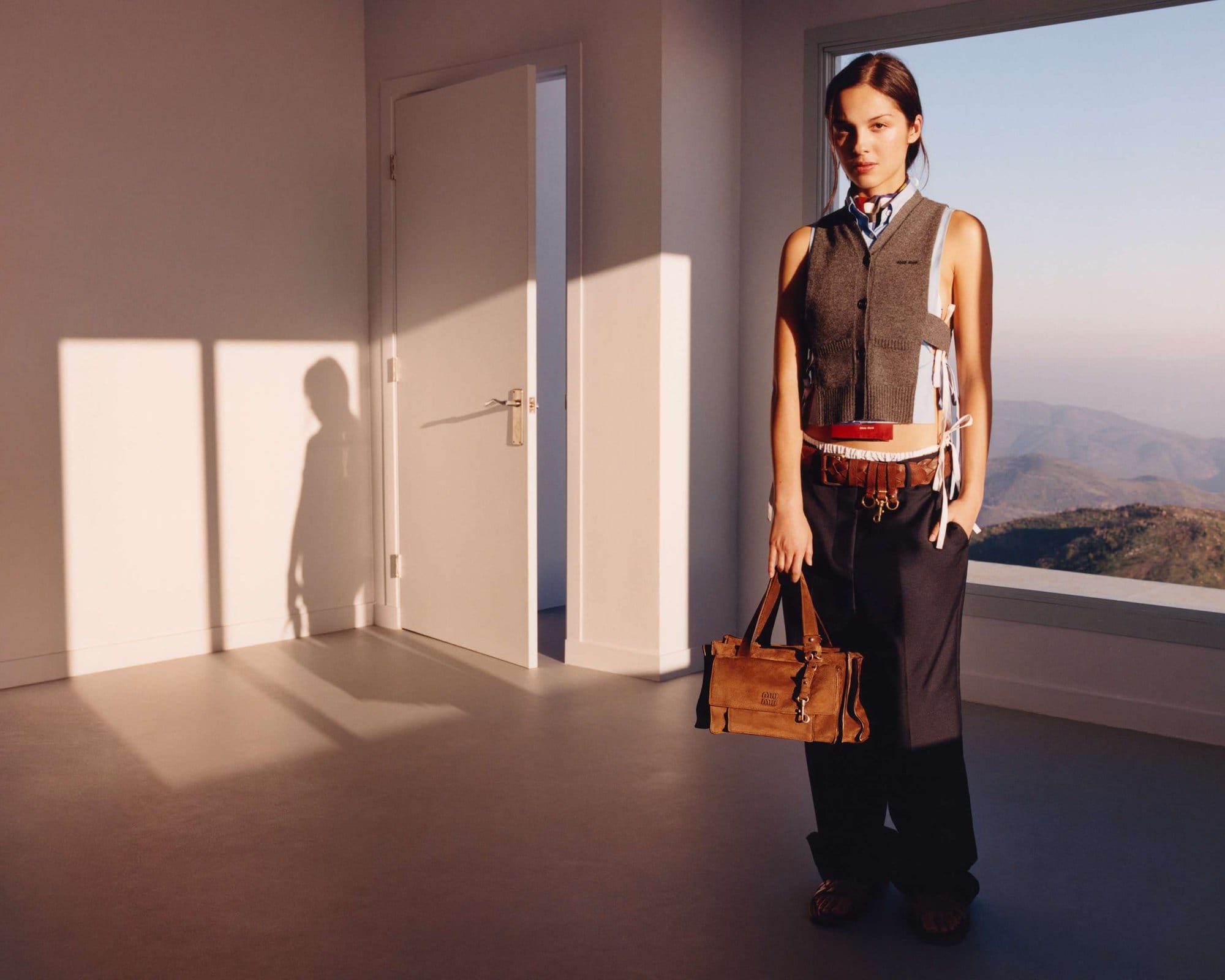 Young woman in vest and trousers holding a tan purse stands in an empty white room with landscape visible through a window.