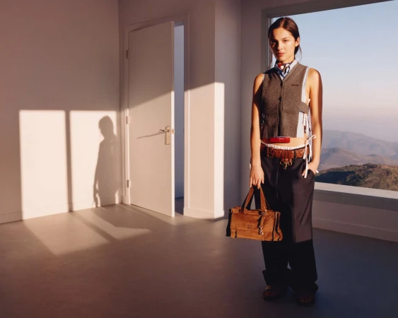 Young woman in vest and trousers holding a tan purse stands in an empty white room with landscape visible through a window.