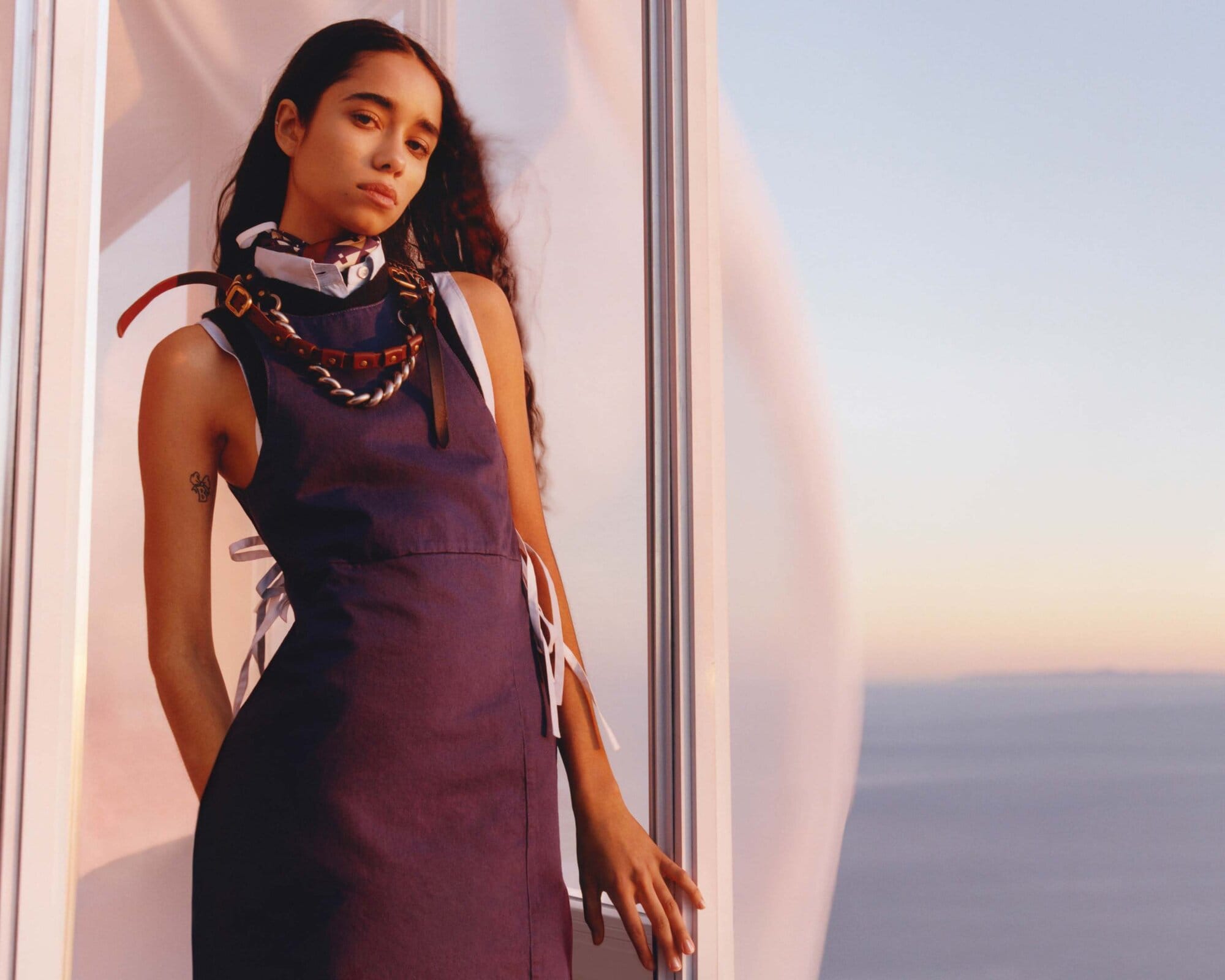 Woman in a navy dress poses in a naturally lit space with large windows showcasing the sky with a horizon, necklace detail.