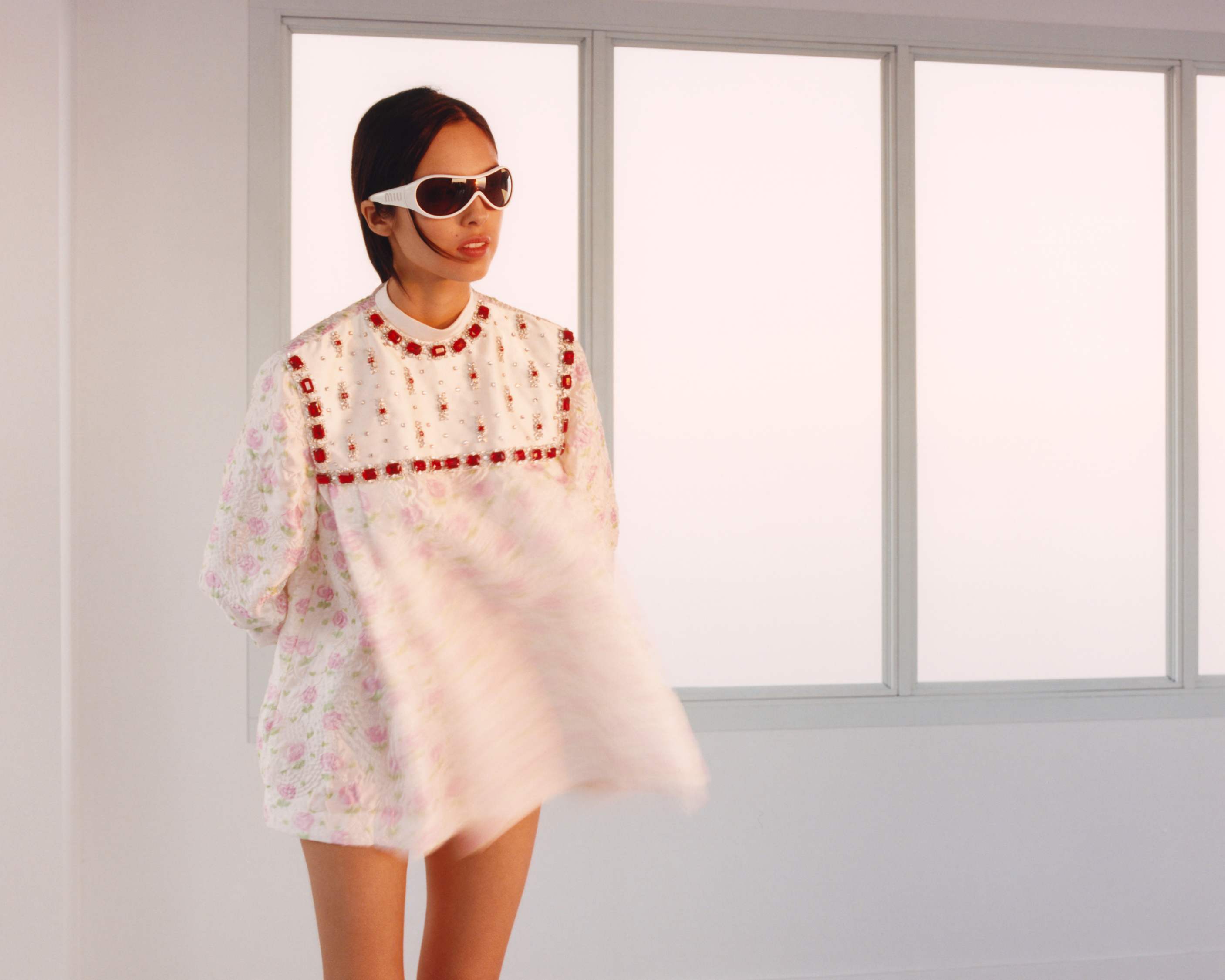 Model wearing white sunglasses and patterned dress in front of large pane windows for Miu Miu Spring 2026 campaign.