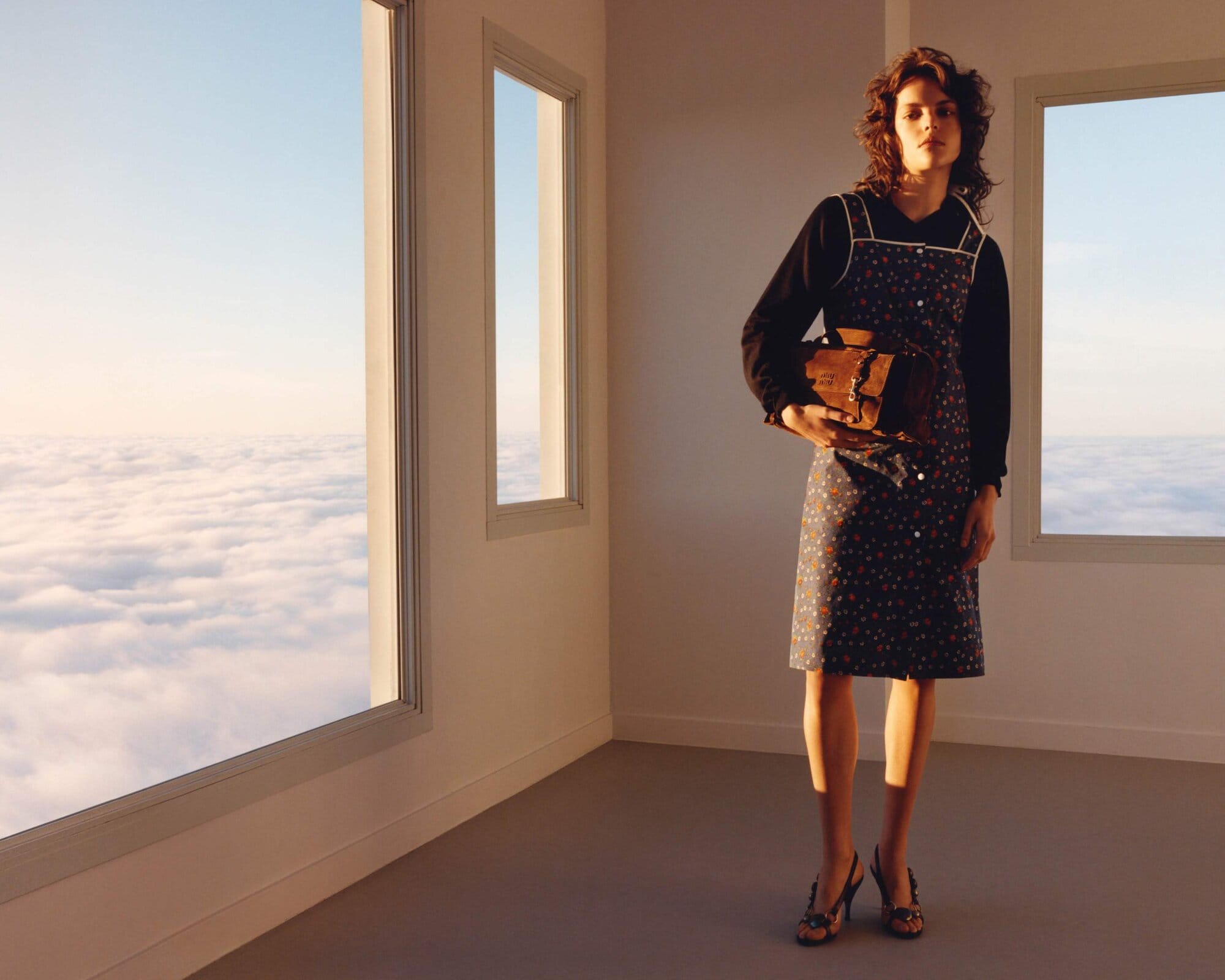A woman in a dress stands in a room with three windows revealing clouds and blue sky while she holds a brown bag.