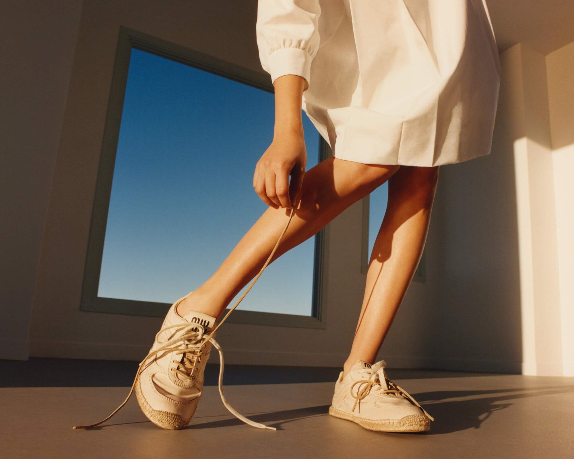 A person ties the laces on a pair of off-white Miu Miu sneakers in a bright room, with a large window in the background showing a blue sky.
