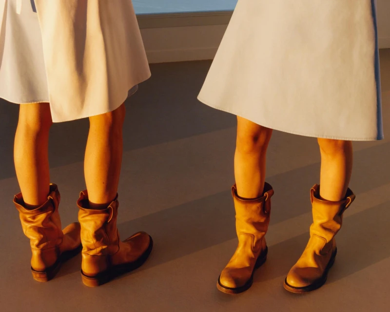 Two models pose in fashionable brown leather boots and white dresses, casting a warm shadow in a minimalist indoor fashion shot.