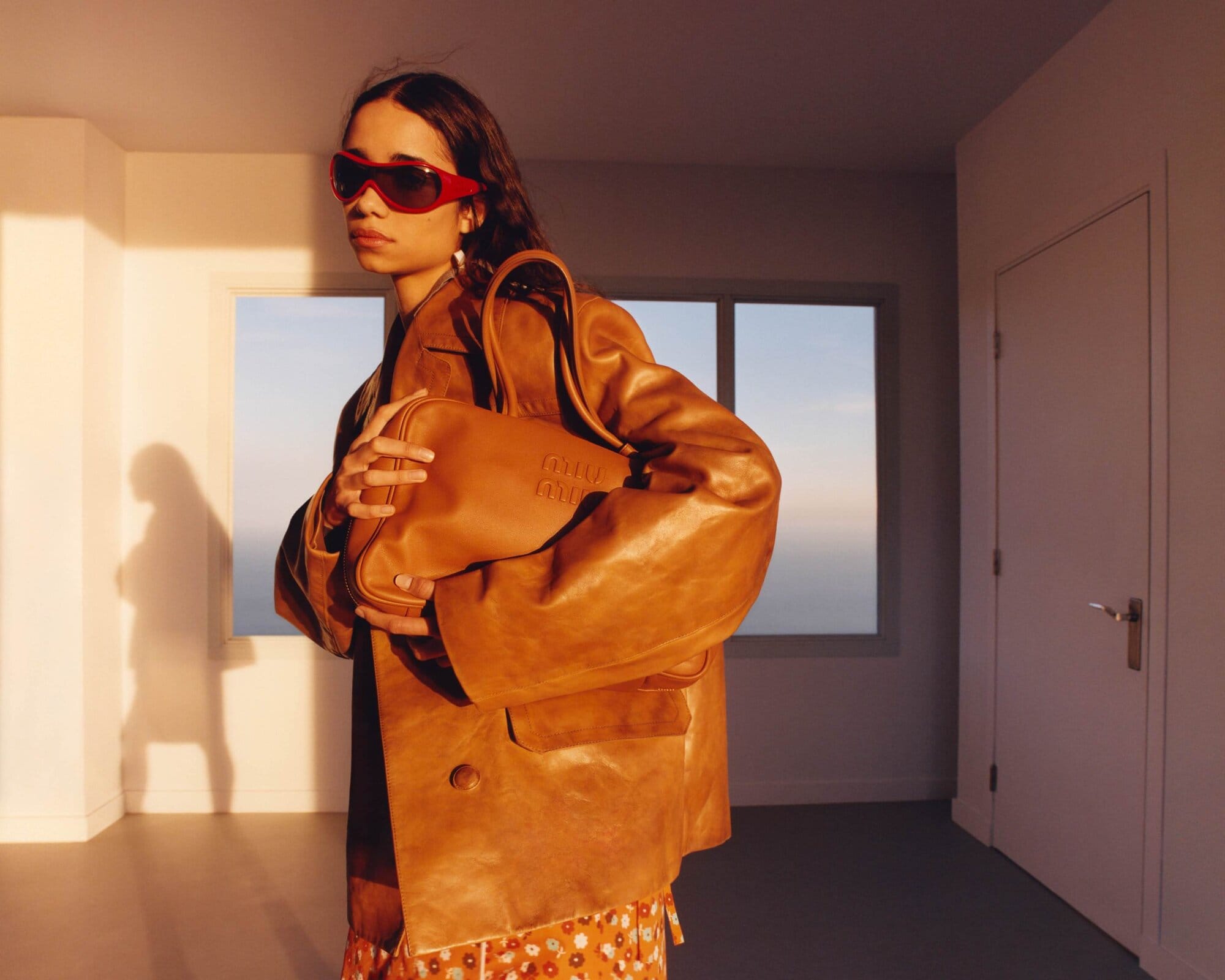 Indoor fashion shot featuring a woman in a tan jacket holding a tan purse, wearing red sunglasses in a minimalist room setting.