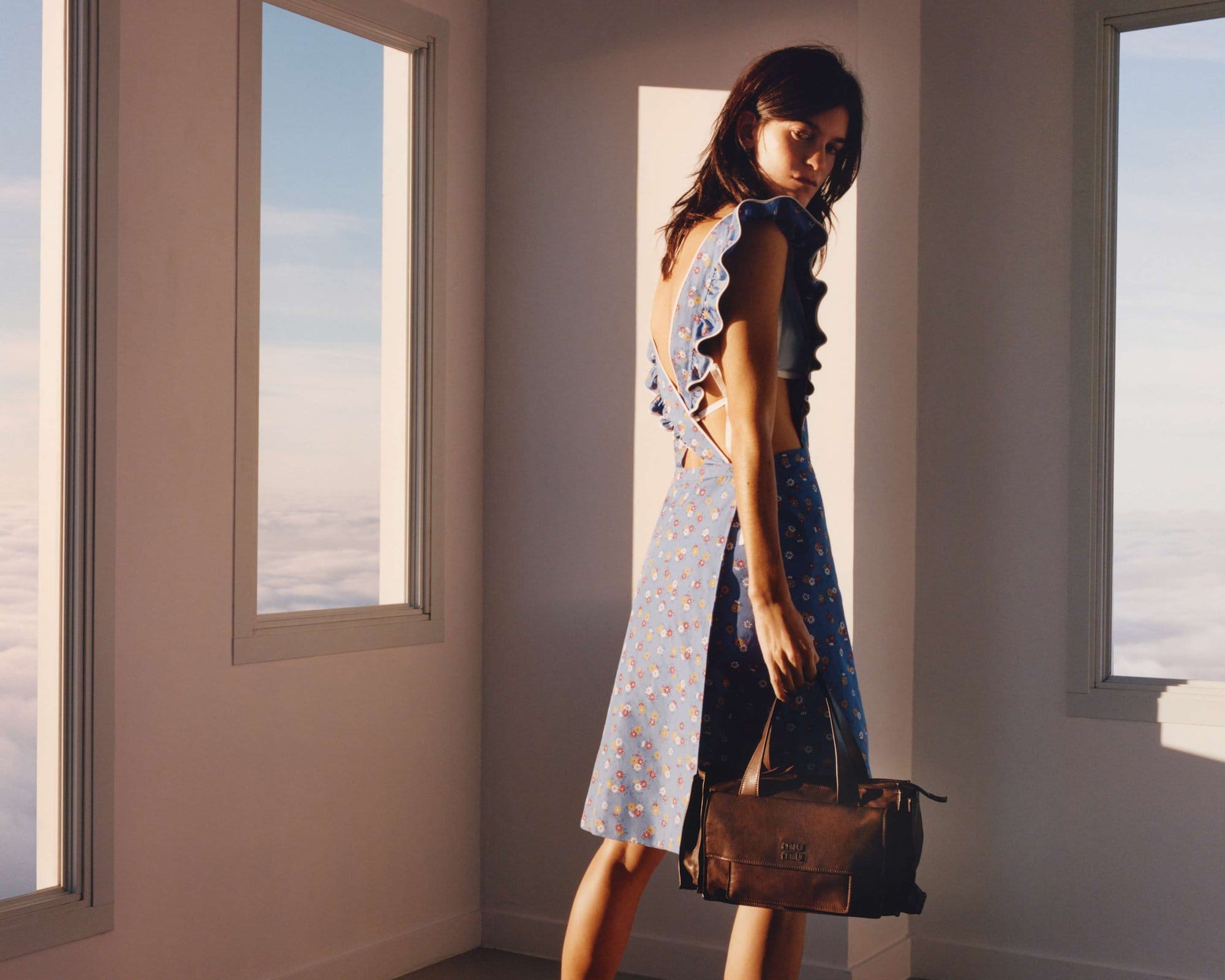 Woman in a blue floral dress with ruffles stands by a window overlooking clouds, holding a brown leather satchel bag.