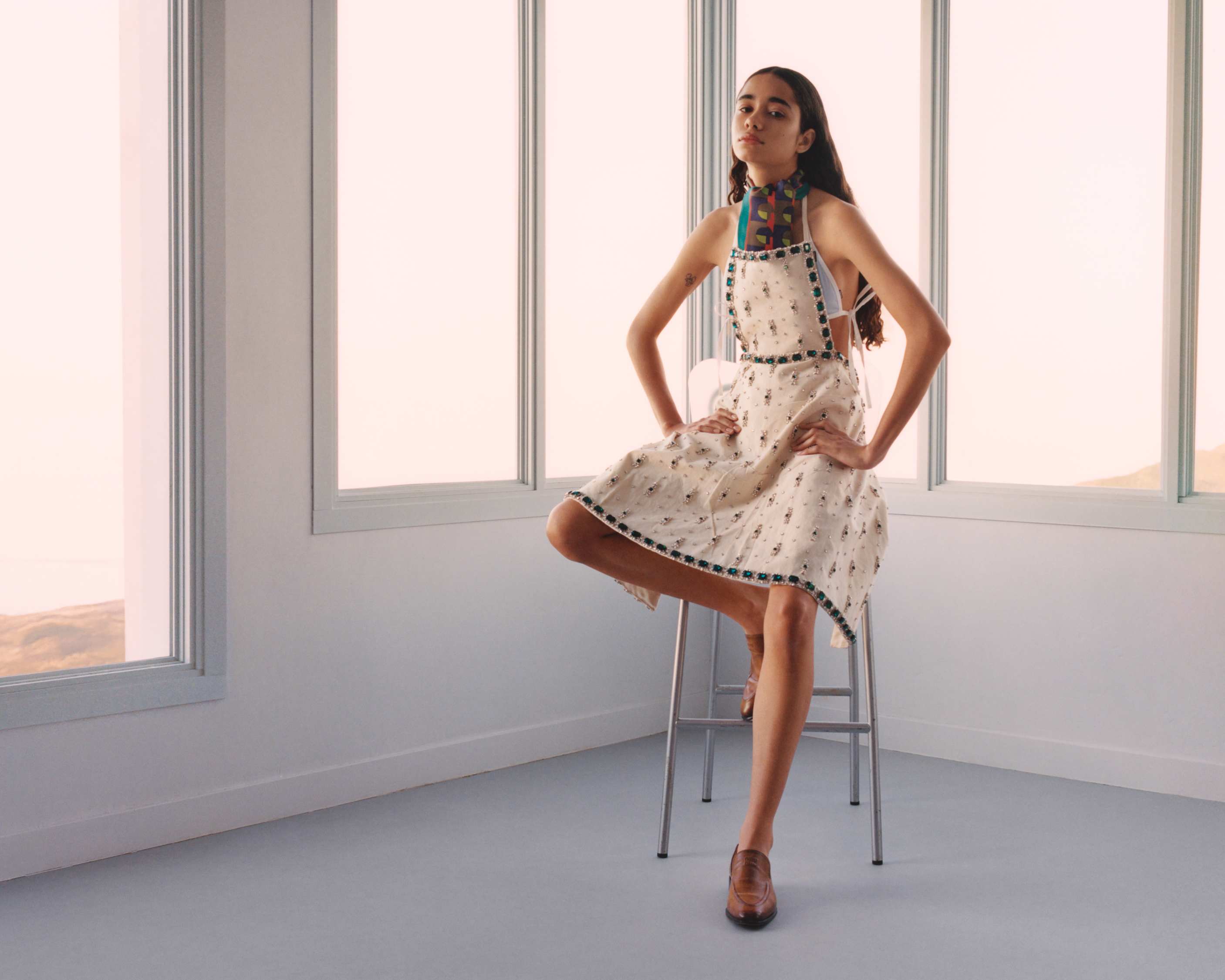Model wearing patterned apron dress seated on high stool against bright windows