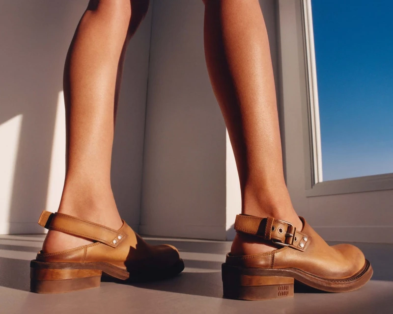 Woman walking in brown leather platform shoes against a blue sky near a white wall in bright sunlight and strong shadows.