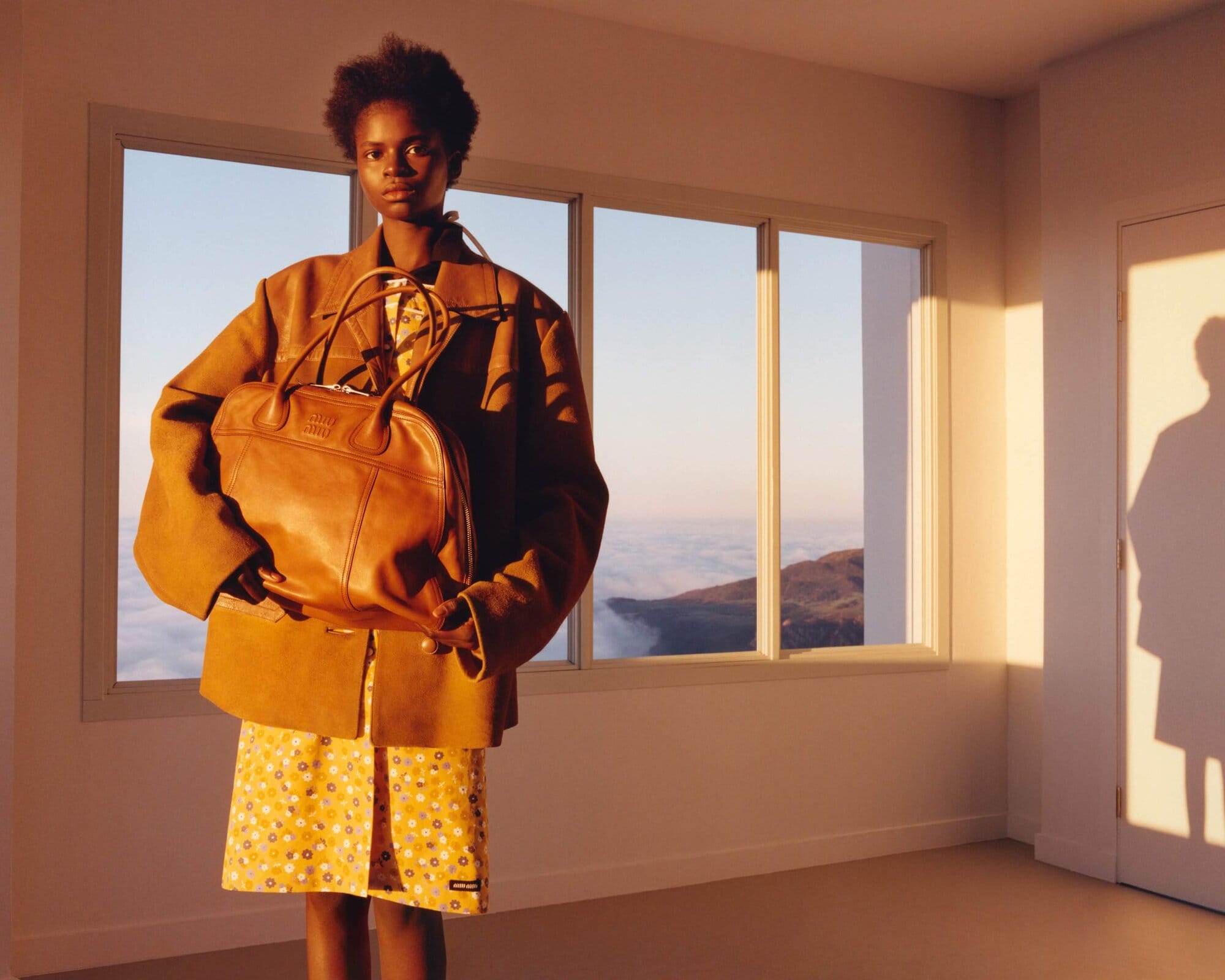 Woman in tan jacket holds brown leather purse inside a sunlit room with a mountain view from the window, creating a fashion editorial.