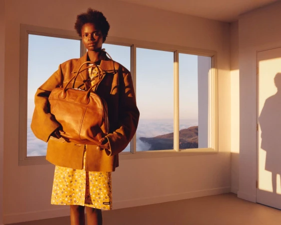 Woman in tan jacket holds brown leather purse inside a sunlit room with a mountain view from the window, creating a fashion editorial.