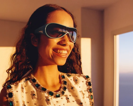 Smiling woman wearing a beaded white top and blue sunglasses standing near a window with outdoor scenic background, bathed in warm light.