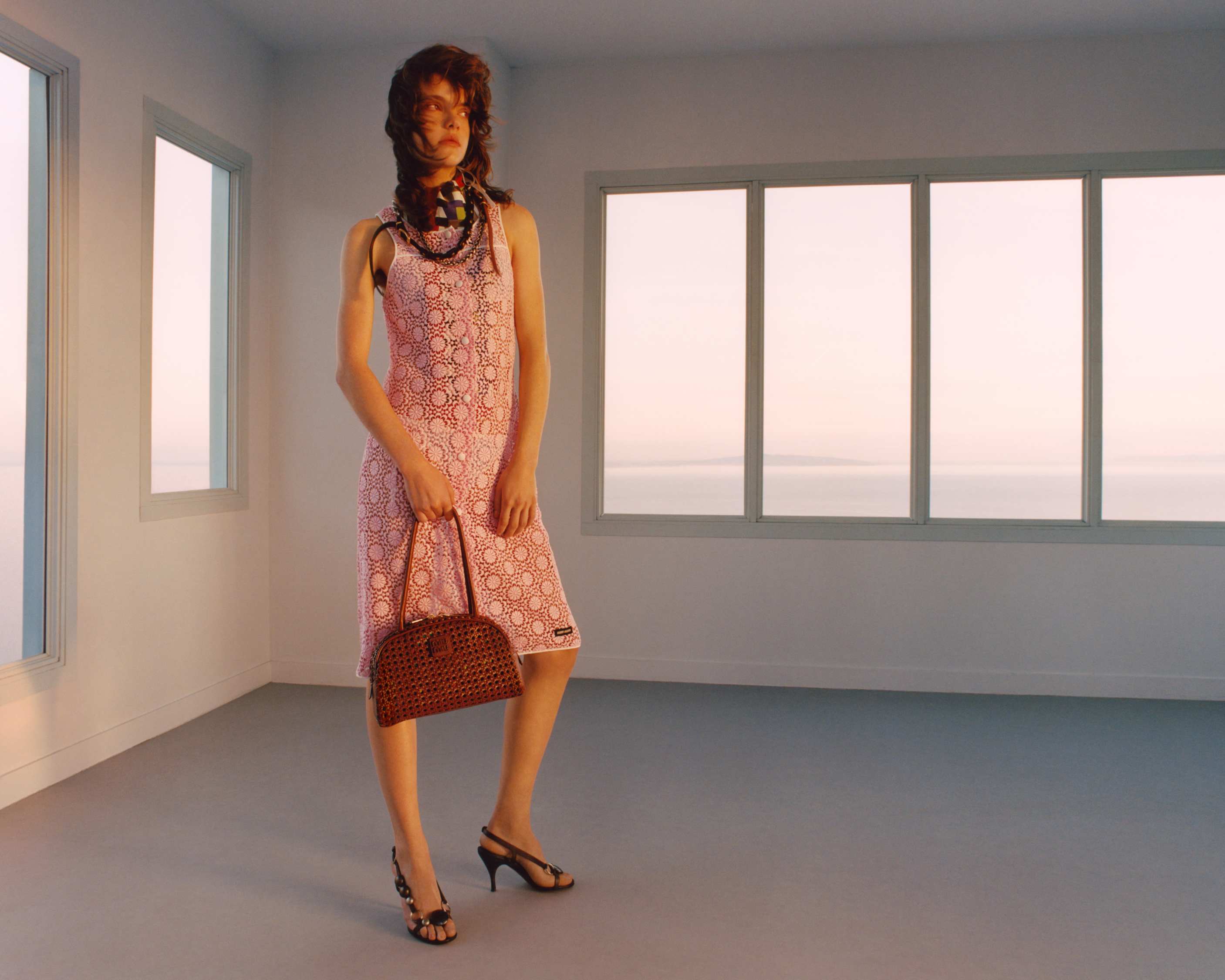 A brunette female fashion model poses in a pink patterned dress with a brown purse, set against a backdrop of windows and soft lighting.