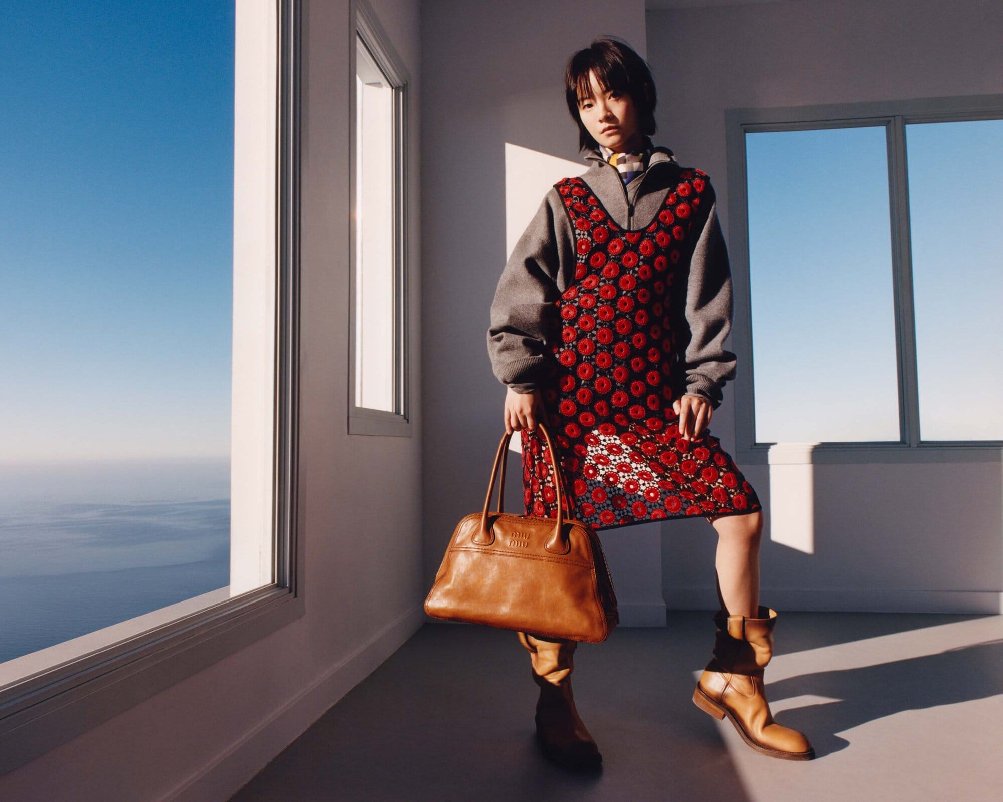 Indoor studio shot of a stylish Asian woman in a floral dress, grey sweater, and tan boots, holding a leather bag near a large window.