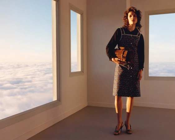 Model standing in a minimal room with large windows overlooking clouds, wearing a patterned dress and holding a brown handbag.
