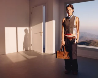 Model standing in a modern interior holding a brown suede handbag, wearing a grey vest and trousers