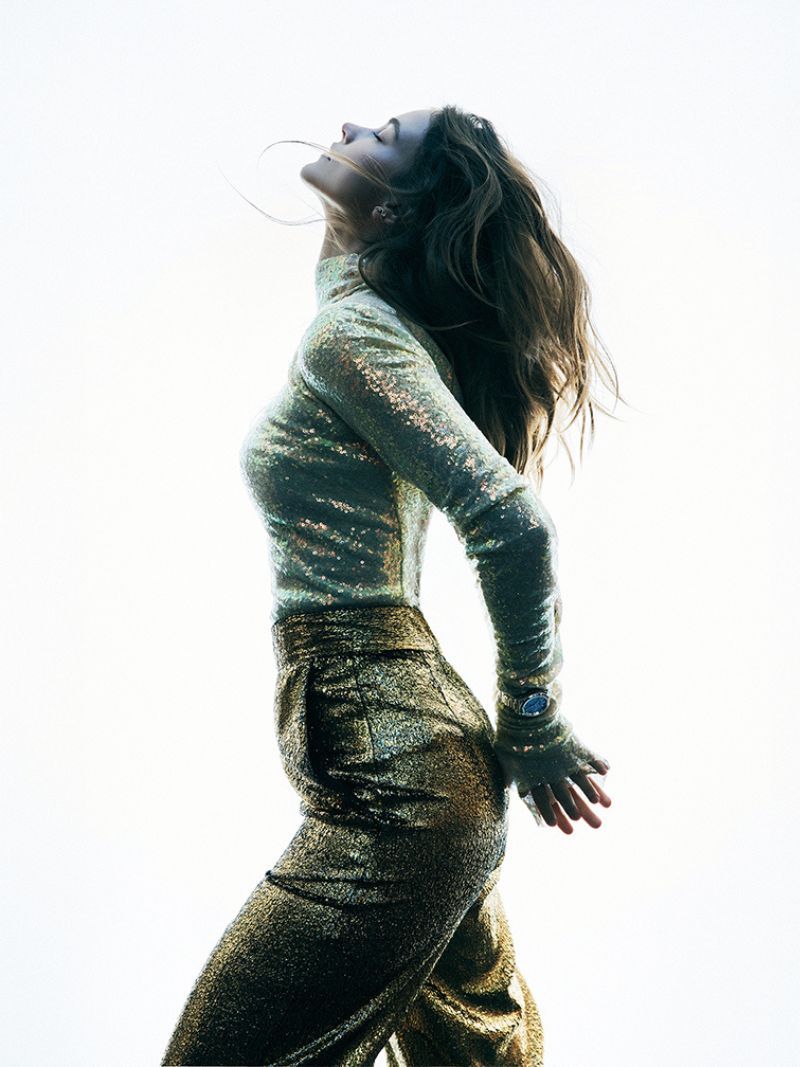 Woman in a shimmering gold outfit stands with her head tilted back in a profile shot against a bright, neutral background.