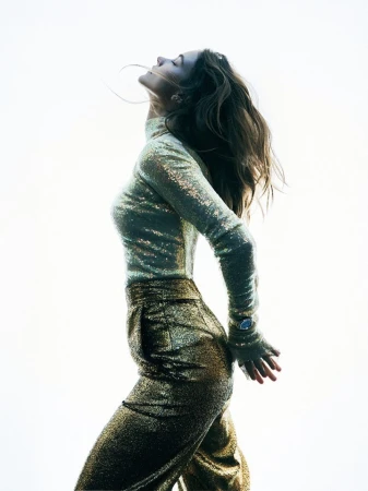 Woman in a shimmering gold outfit stands with her head tilted back in a profile shot against a bright, neutral background.