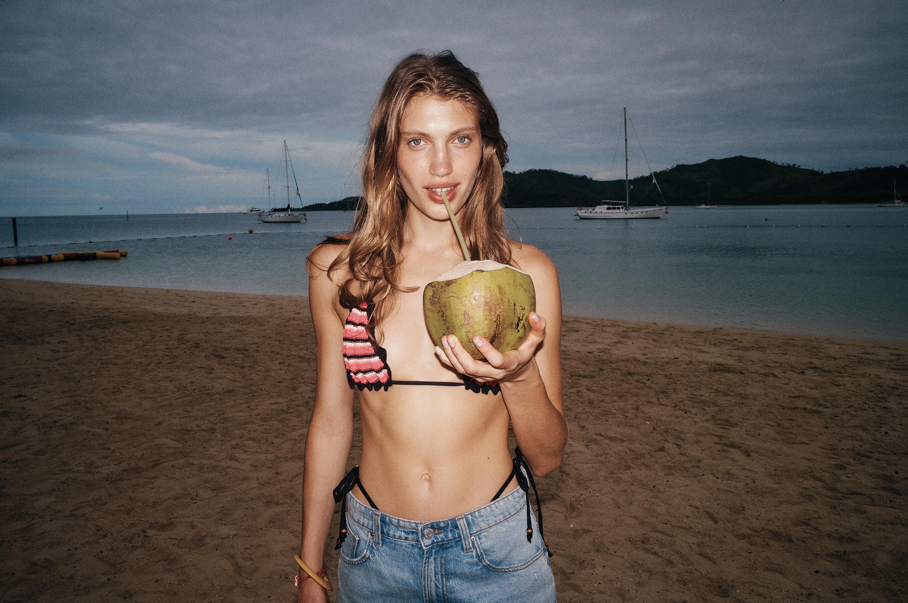 Woman in bikini top and denim shorts poses on a beach, drinking from a coconut with yachts on the water in the background.