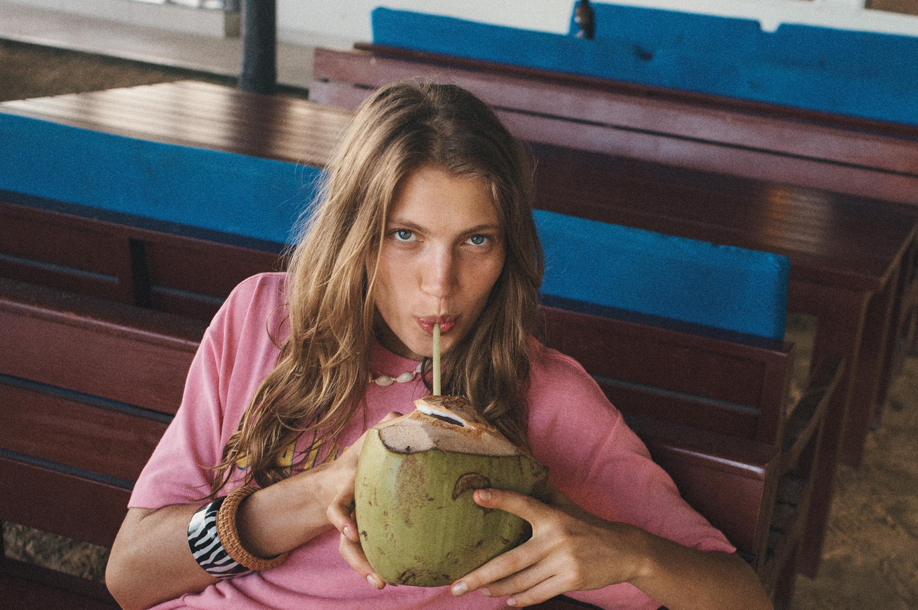 Woman in pink top drinking coconut water with straw at outdoor cafe with wooden tables and benches, shell necklace and bracelets.