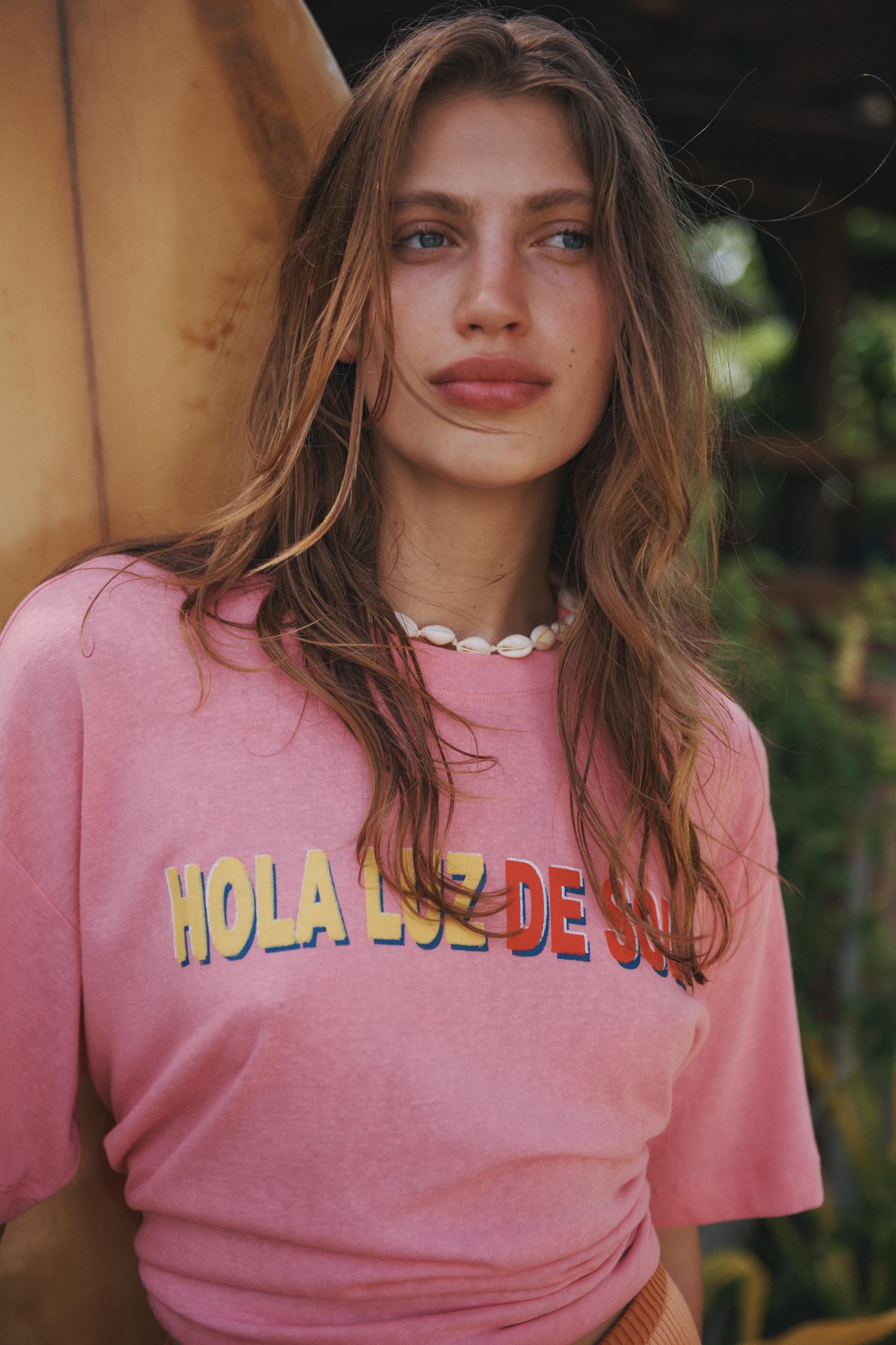 Young woman with long wavy hair poses in front of a surfboard, wearing a pink t-shirt and seashell necklace in an outdoor setting.