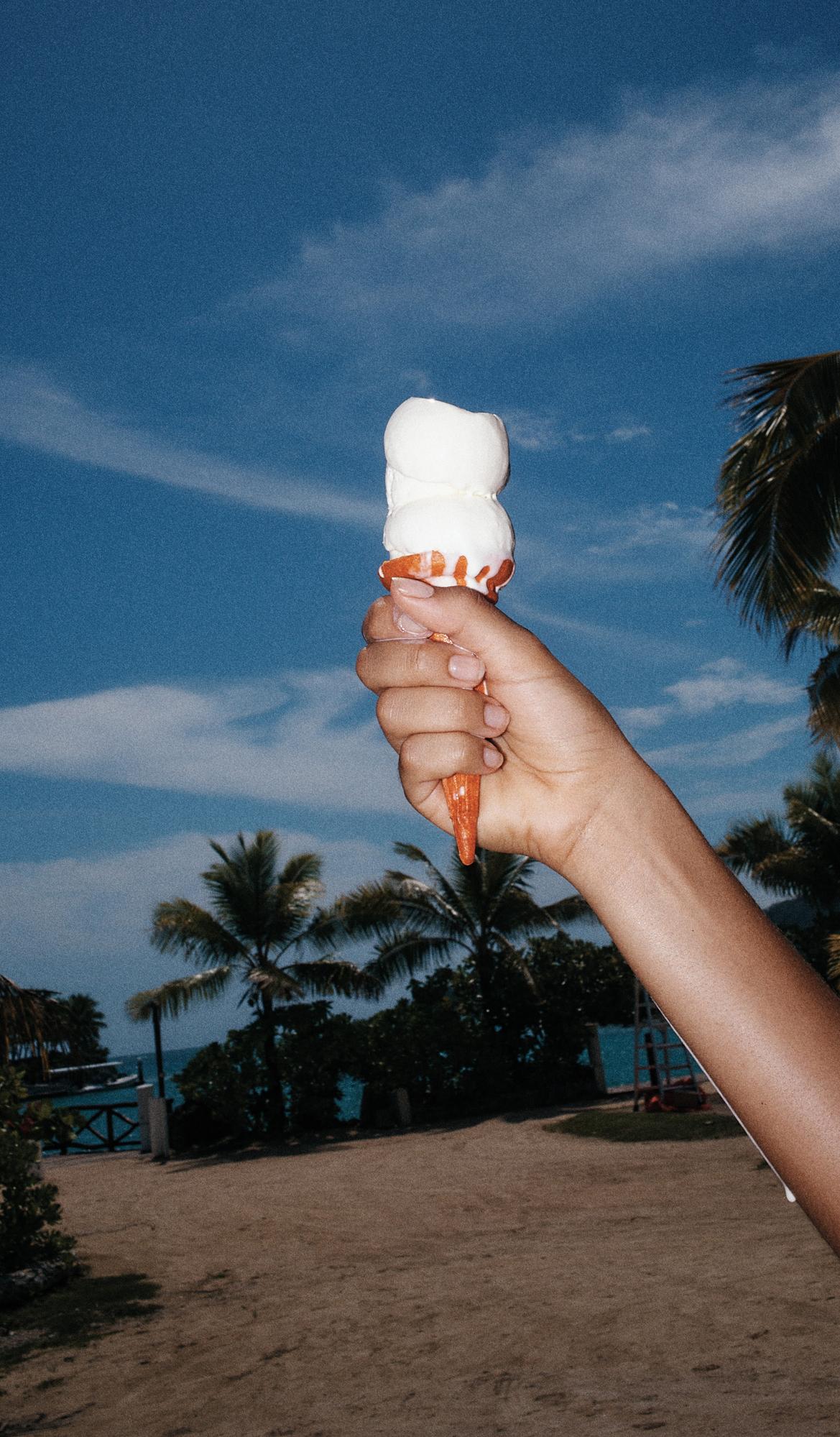 Hand holding a double scoop of ice cream in a cone on a beach with palm trees, blue sky and clouds in the background on a sunny day.