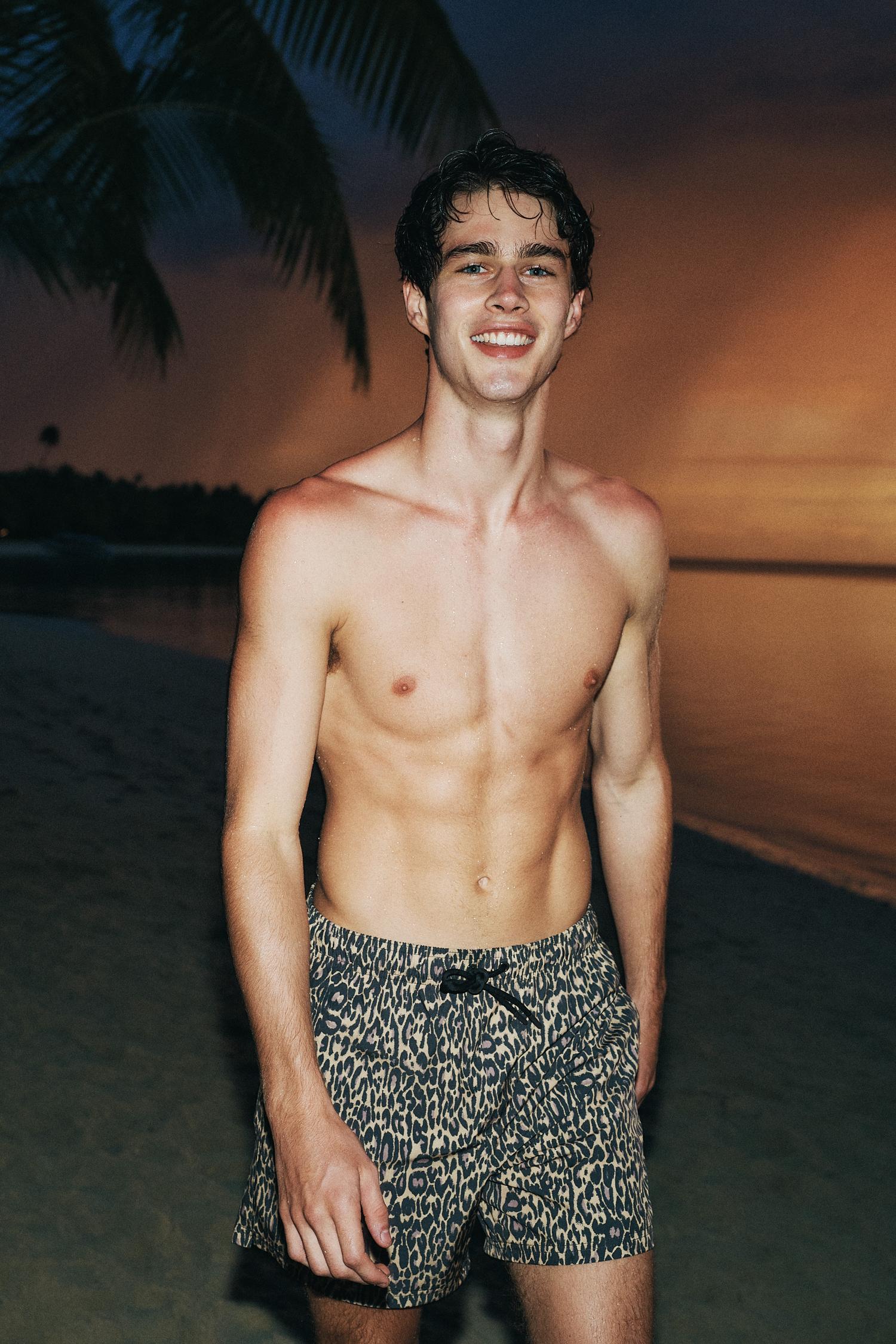 Young man with an athletic build and a smile, posing shirtless in animal print shorts on a sandy beach with a sunset background.