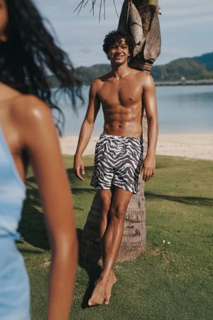 Man poses shirtless in zebra print swim trunks, leaning against a palm tree on a tropical beach with partial view of woman.