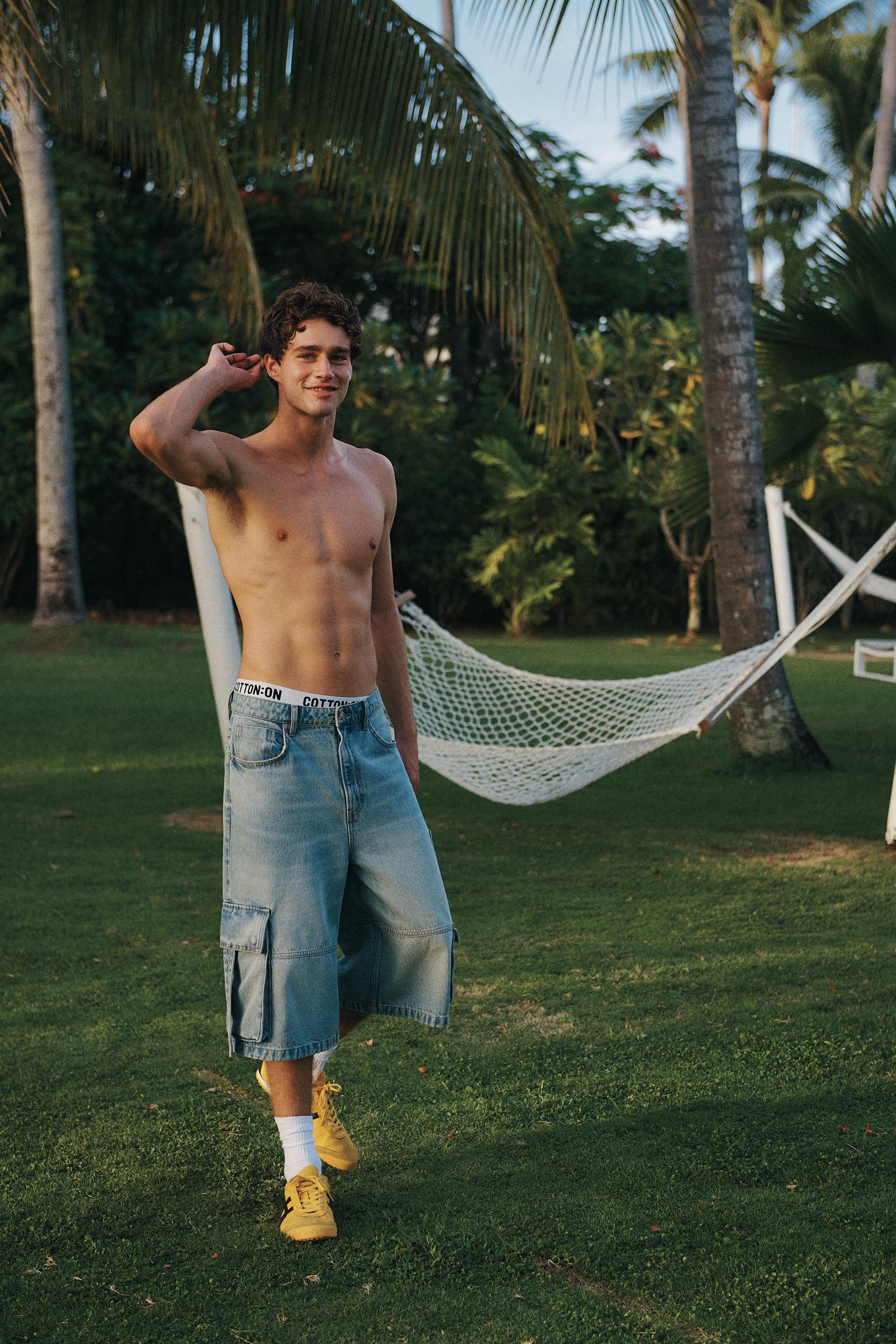 A muscular shirtless man poses outdoors on a grassy lawn wearing denim culottes, yellow sneakers, and white socks, with palm trees behind.