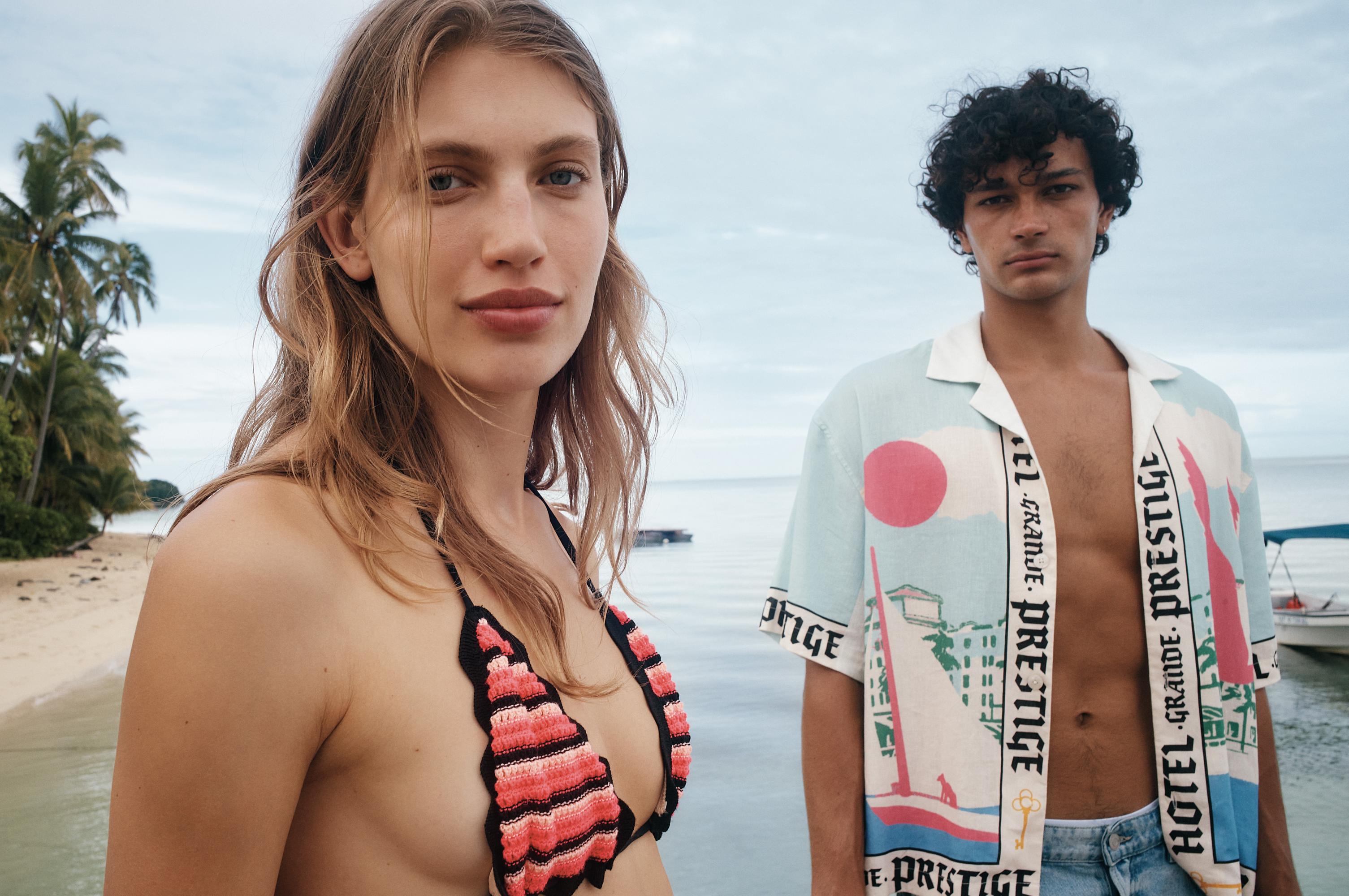 Two models, a blonde woman in a bikini and a curly-haired man in a patterned shirt, pose on a tropical beach with palm trees and a boat.