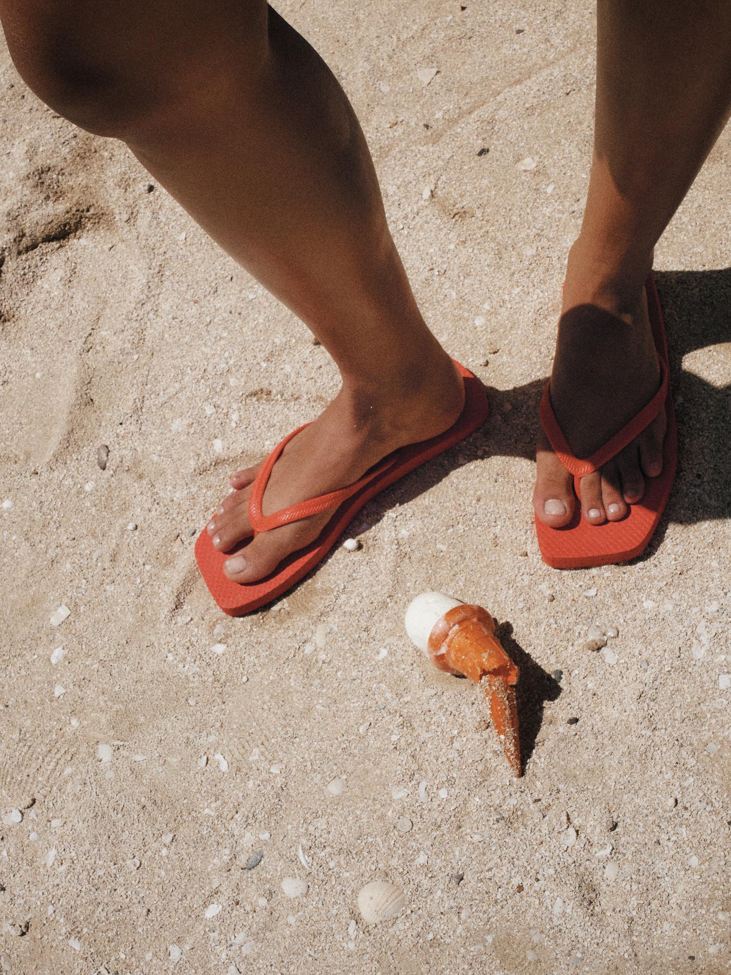 Person standing on sand near partially eaten ice cream cone wearing orange flip flops on a beach during sunny day.