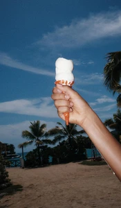Hand holding a melting ice cream cone against a sunny beach background with palm trees under a bright blue sky.