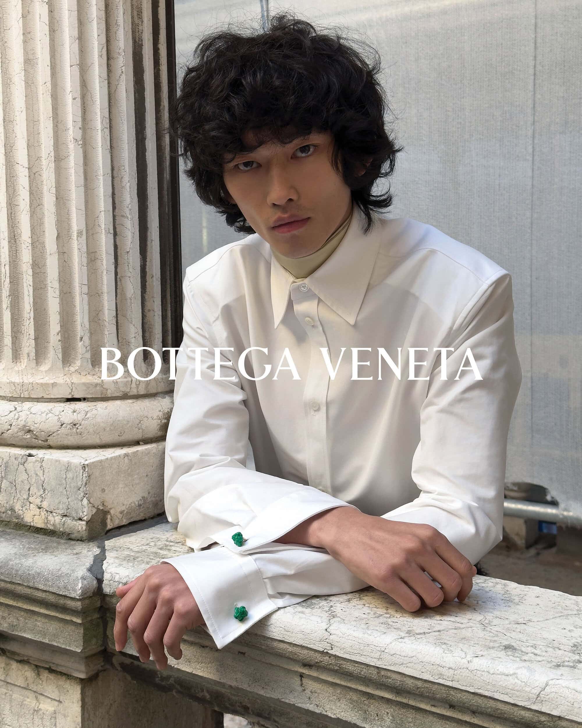 An Asian male fashion model with dark curly hair poses in a white button-down shirt, leaning against a stone wall, showcasing a Bottega Veneta ad.