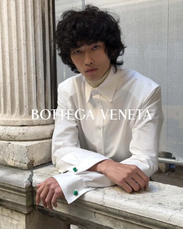 An Asian male fashion model with dark curly hair poses in a white button-down shirt, leaning against a stone wall, showcasing a Bottega Veneta ad.