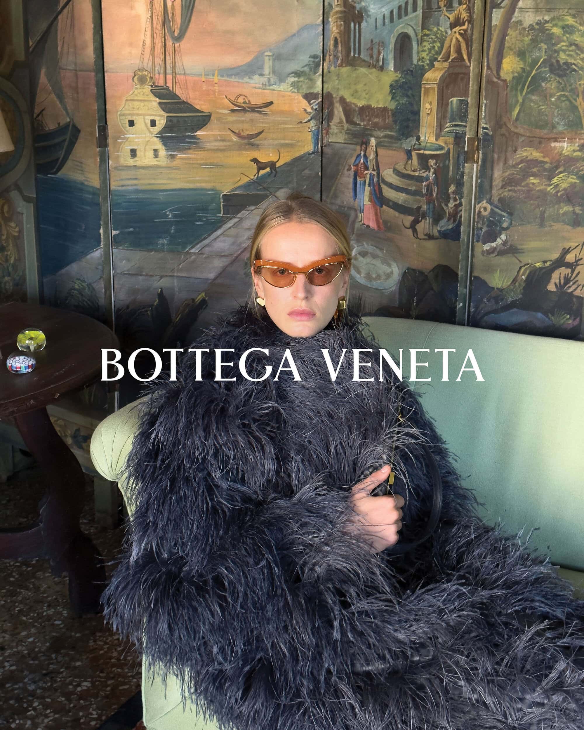 A woman wearing sunglasses and a large feathered coat sits on a couch in front of a painted backdrop with the text BOTTEGA VENETA.