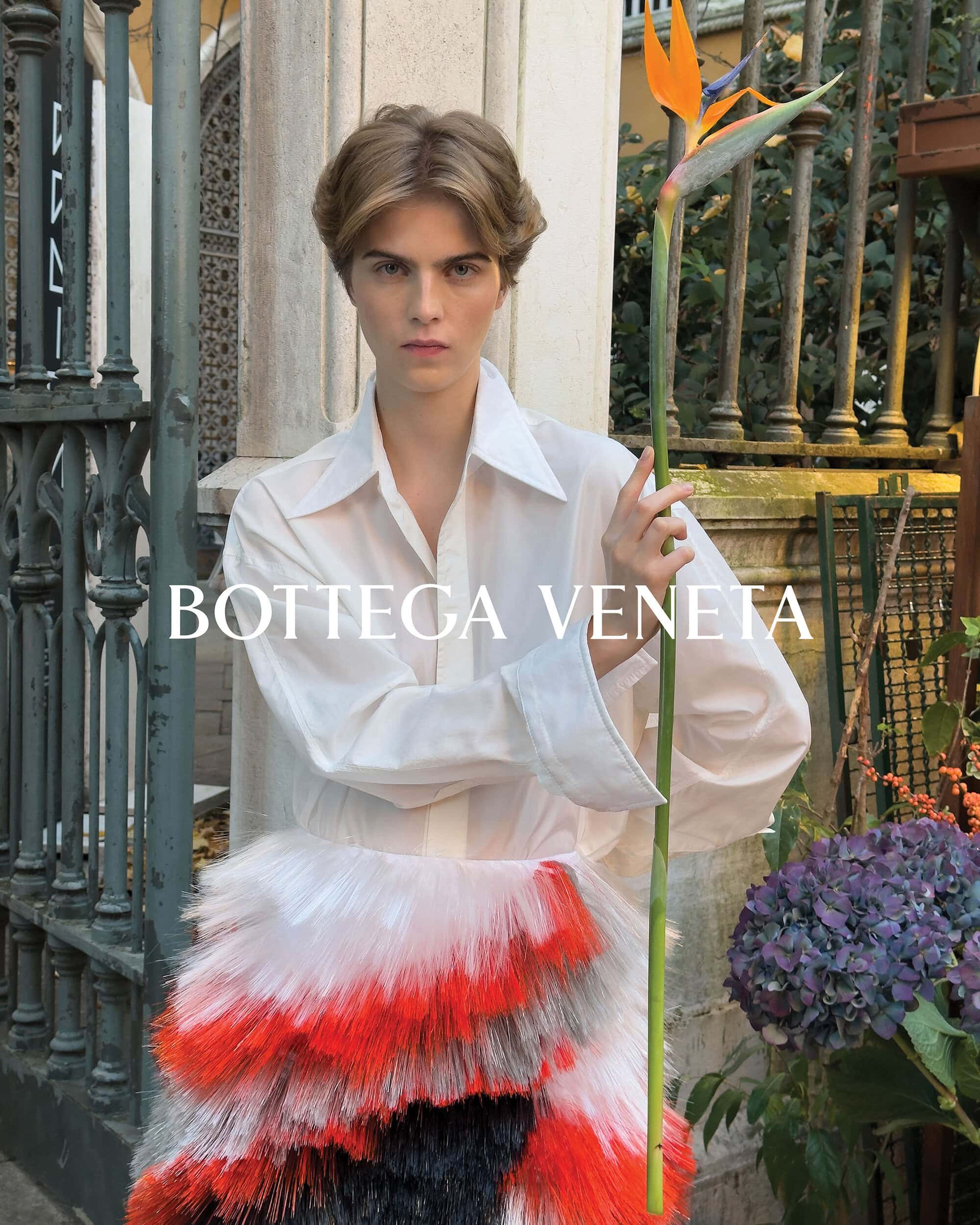 Elegant woman in white shirt and feathered skirt holds a Bird of Paradise flower, posing in front of an iron gate and lush greenery.