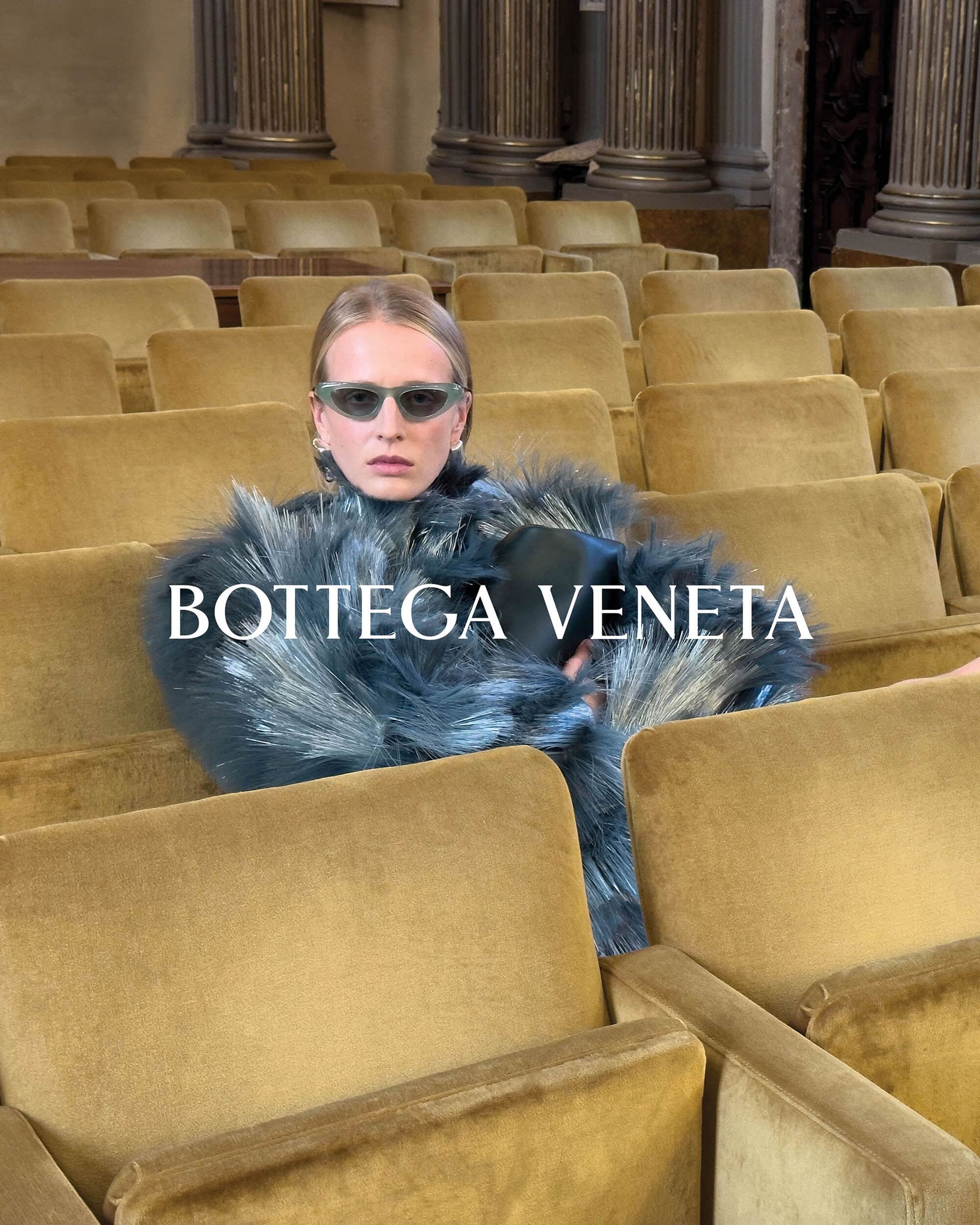 A blonde woman sits in gold theater seats wearing sunglasses and a fur jacket with the BOTTEGA VENETA logo across the center.
