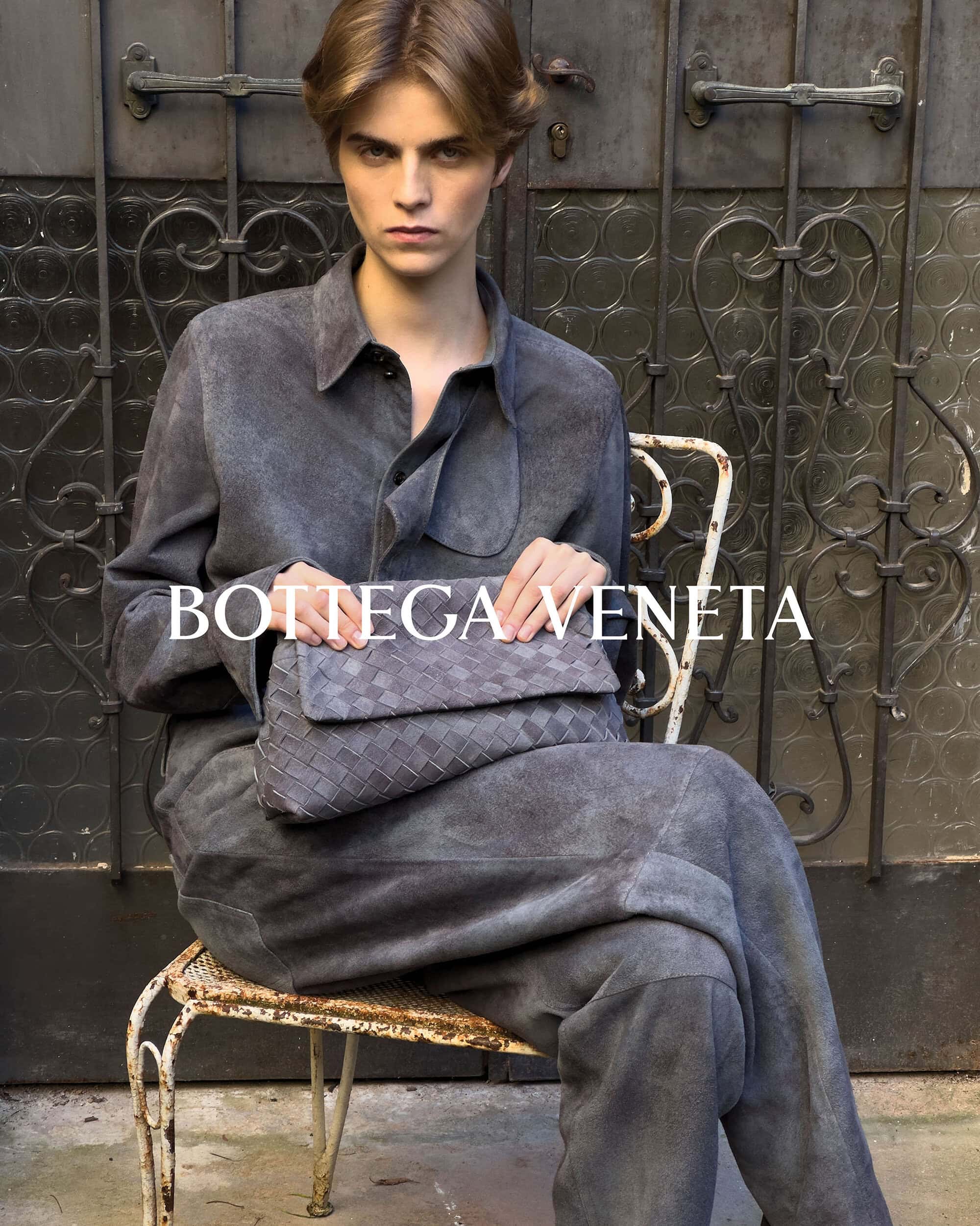 A person with blond hair sits on a white wrought iron chair, holding a grey woven clutch bag, wearing a grey shirt and pants.