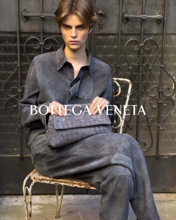 A person with blond hair sits on a white wrought iron chair, holding a grey woven clutch bag, wearing a grey shirt and pants.